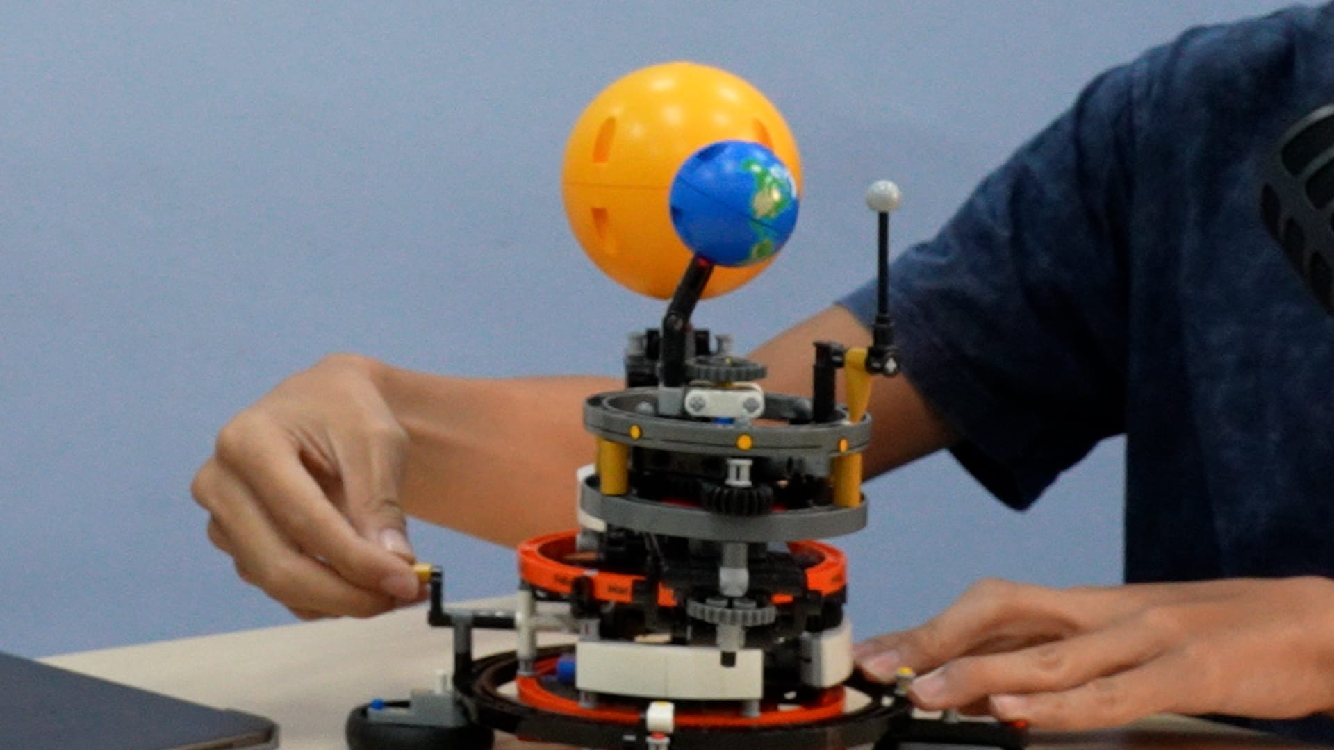 A person adjusts a LEGO orrery model featuring an orange sun, blue Earth, and white moon on a mechanical base.