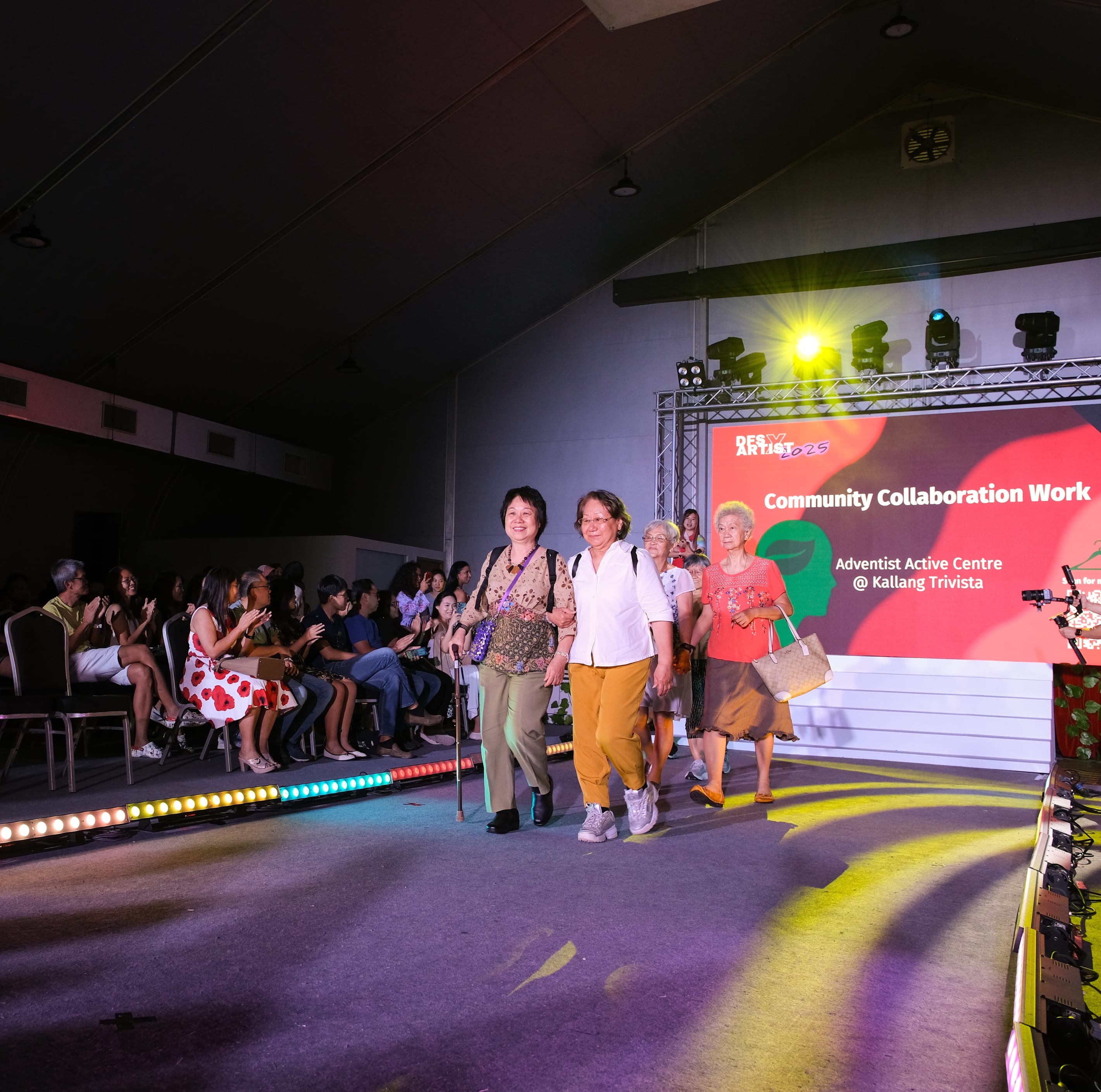 Elderly women walk a lit runway at a community event as seated audience applauds, with a red "Community Collaboration Work" screen behind.
