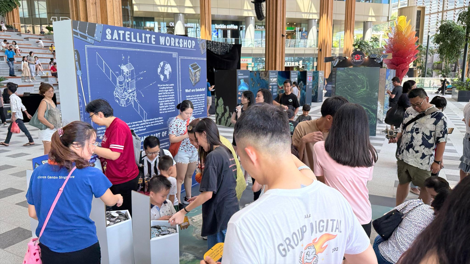 Crowd of adults and children gathered around displays at an outdoor Satellite Workshop event in a mall atrium.