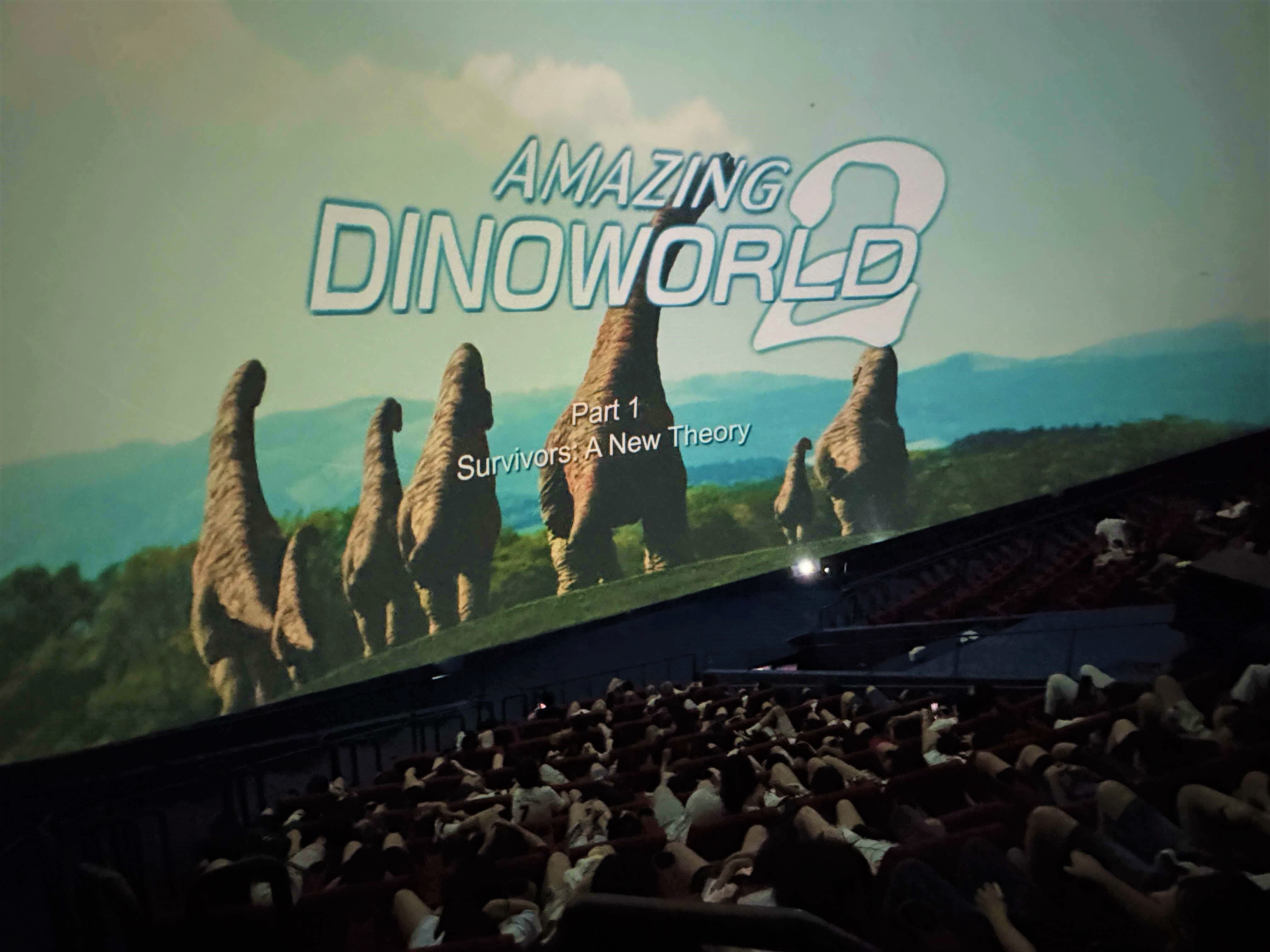 A large screen showing "Amazing Dinoworld 2, Part 1: Survivors: A New Theory" with dinosaurs, viewed by a packed audience.