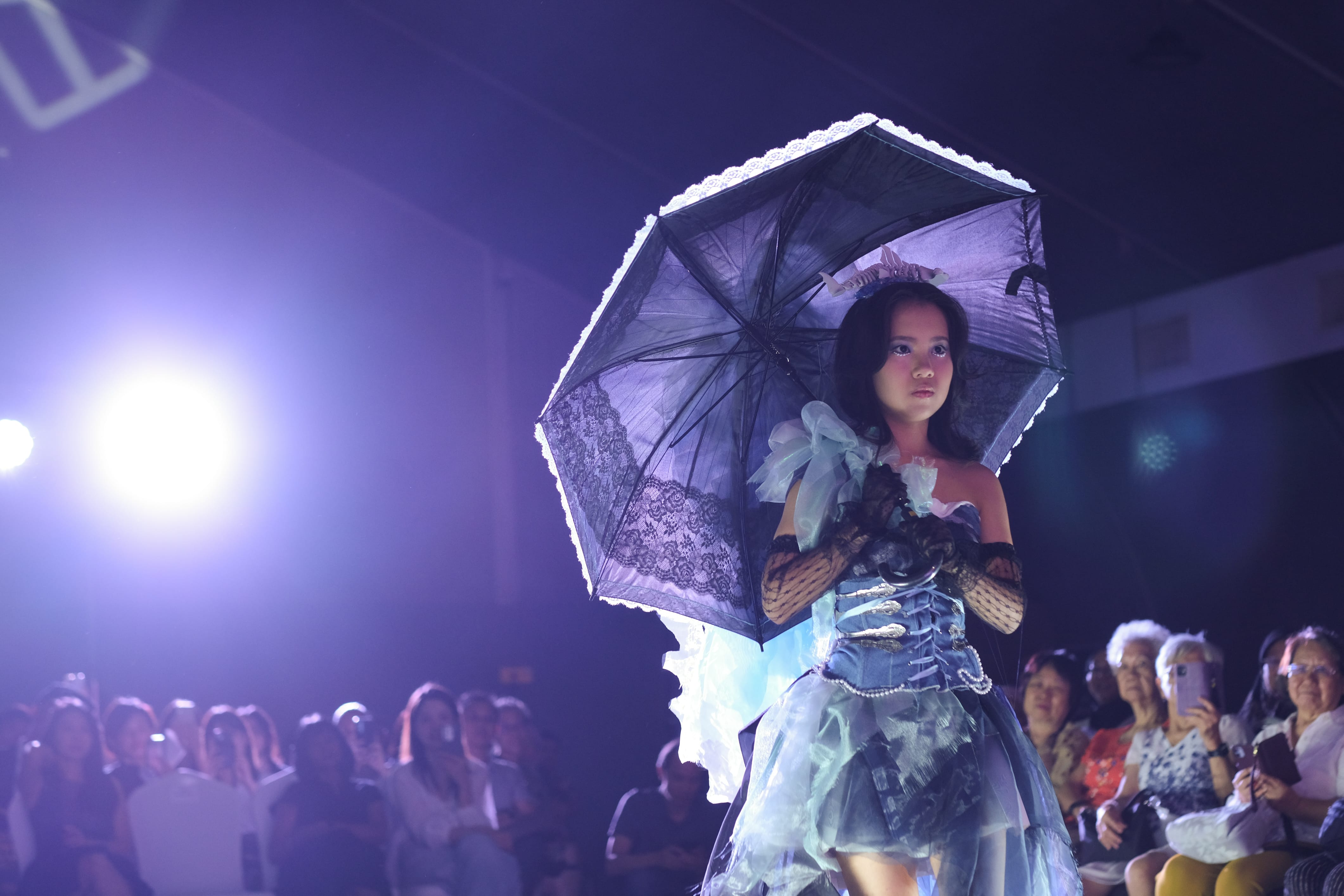 A young woman in a blue lace gown holds a lace parasol on a lit runway with an audience watching.