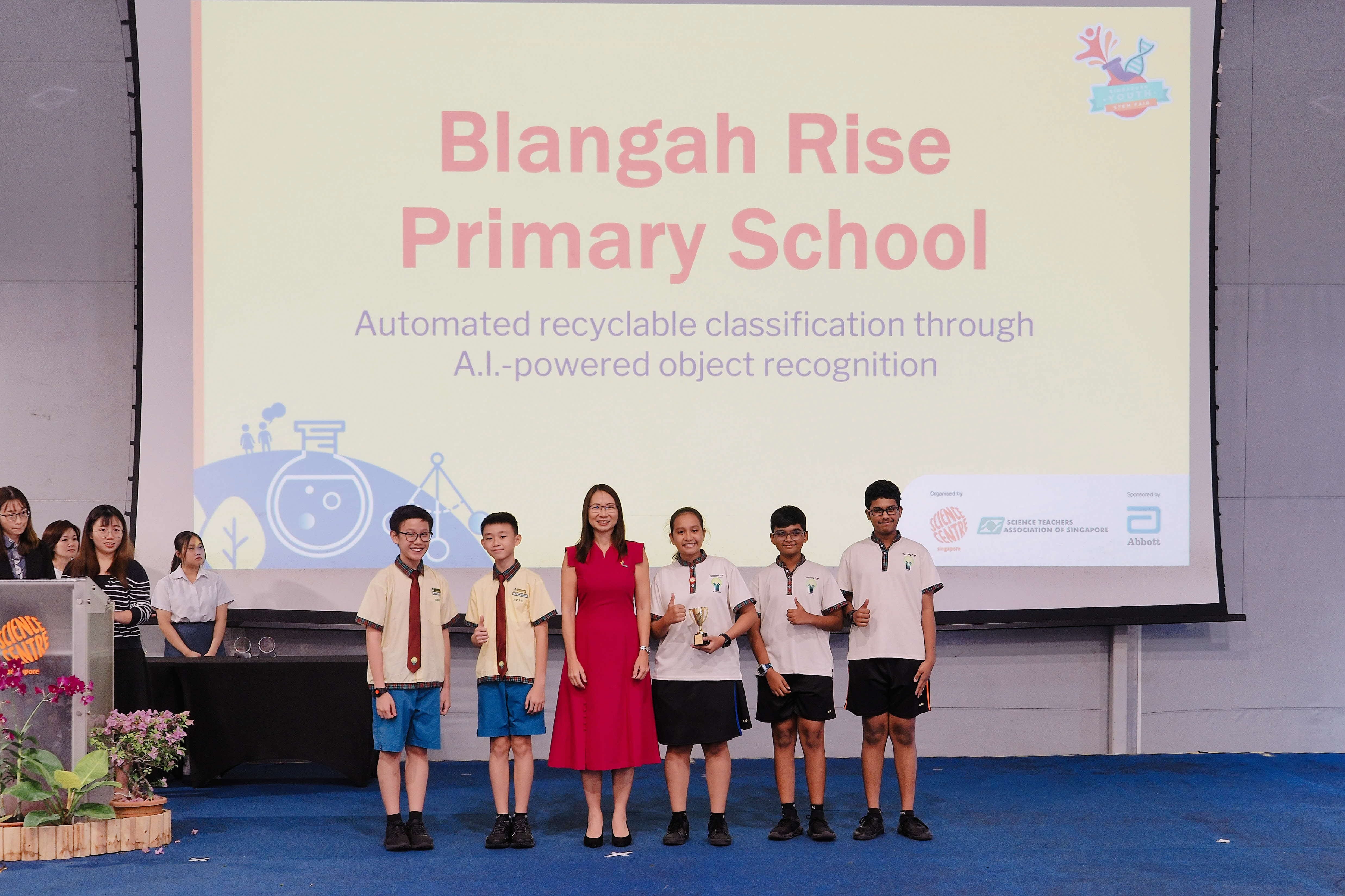 Five students and an adult in red dress pose on stage before a Blangah Rise Primary School presentation slide.