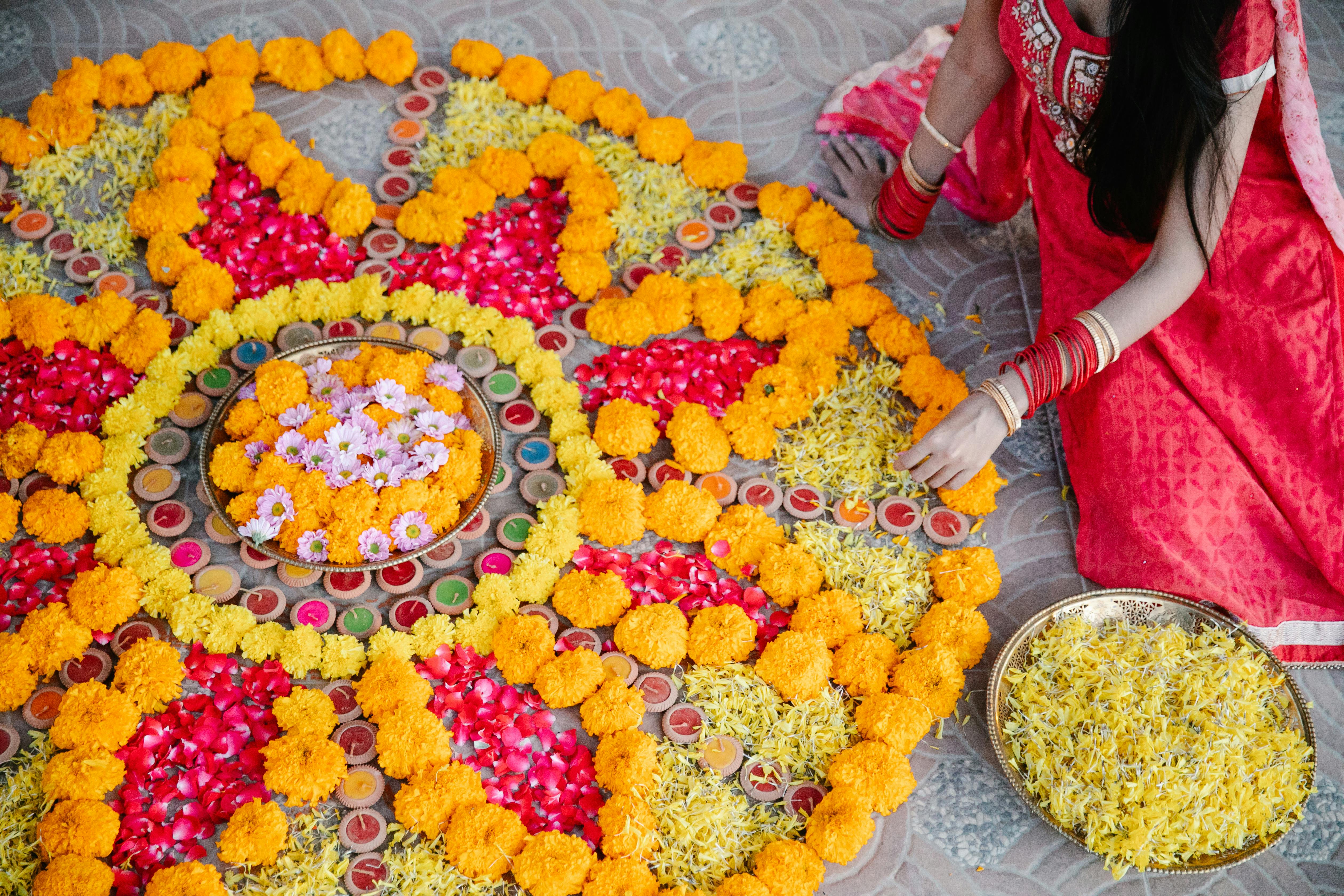 Woman in red saree arranging a colorful flower rangoli with marigolds, rose petals, and small candles on the floor.