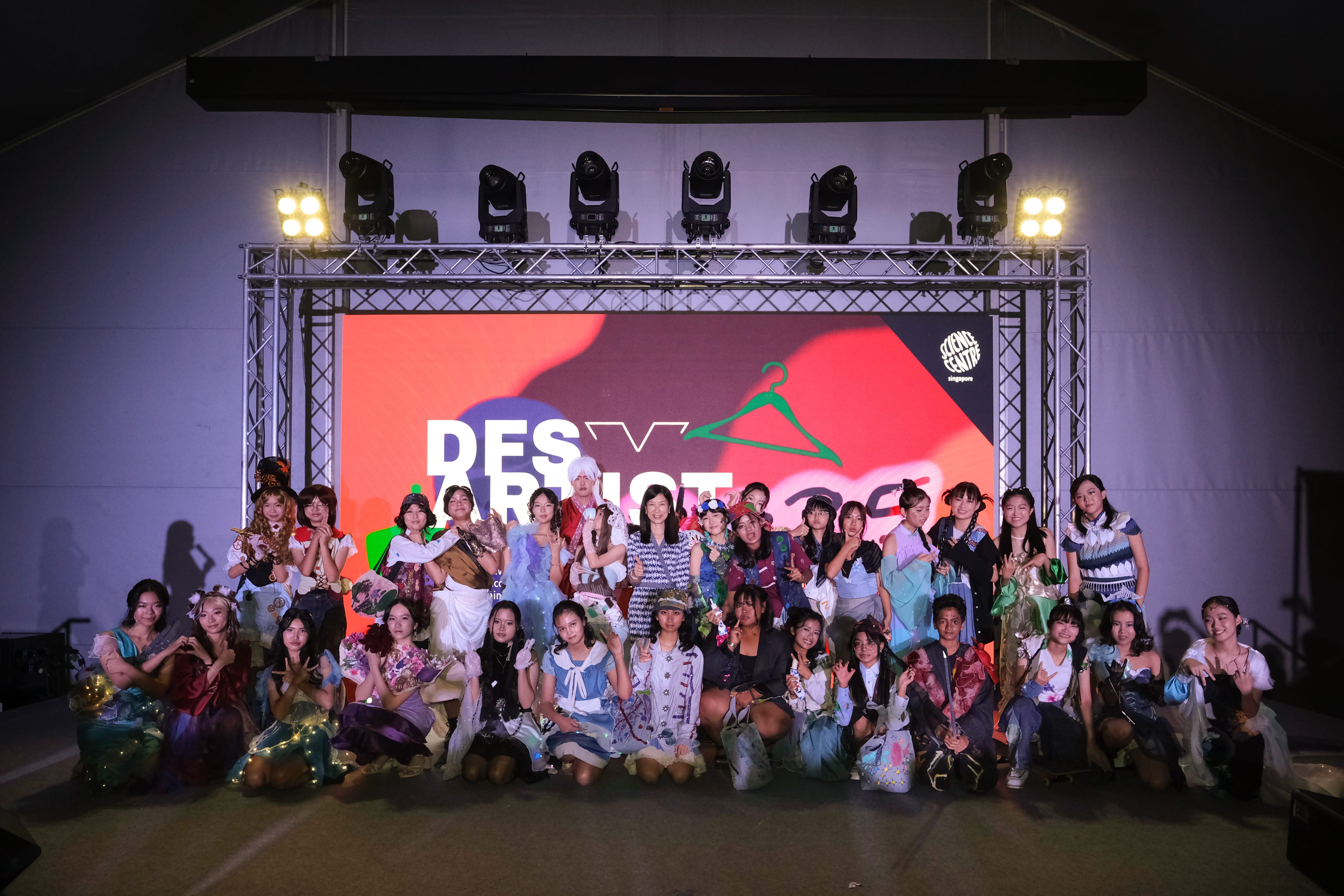 A large group in colorful costumes poses on a stage in front of a red screen reading "DES ARTIST" with stage lights above.