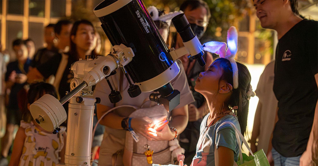 A young girl wearing bunny ears looks into a Sky-Watcher telescope at a nighttime outdoor stargazing event.