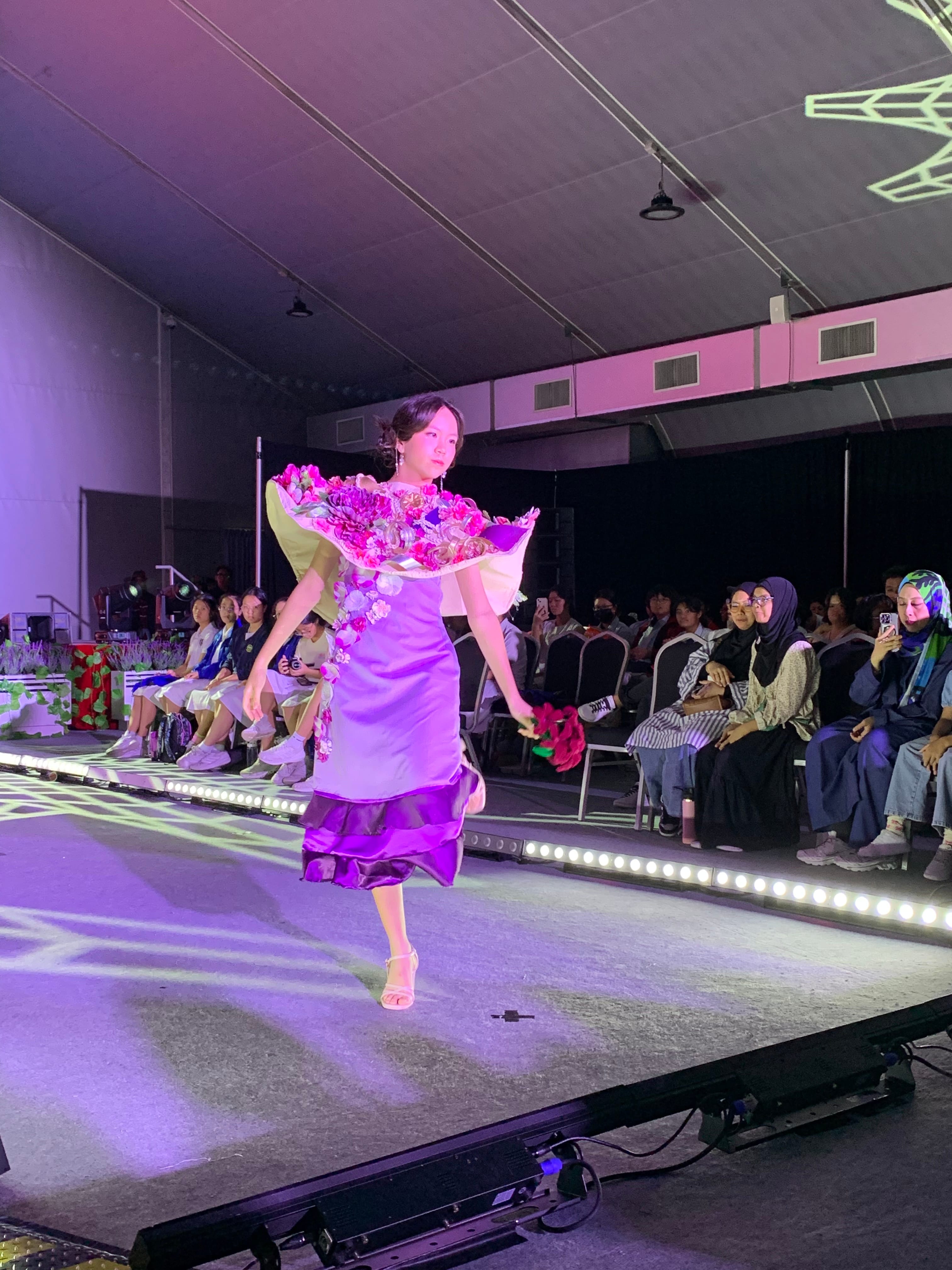 A model walks a lit runway in a purple floral dress with large flower embellishments, as seated audience members watch.