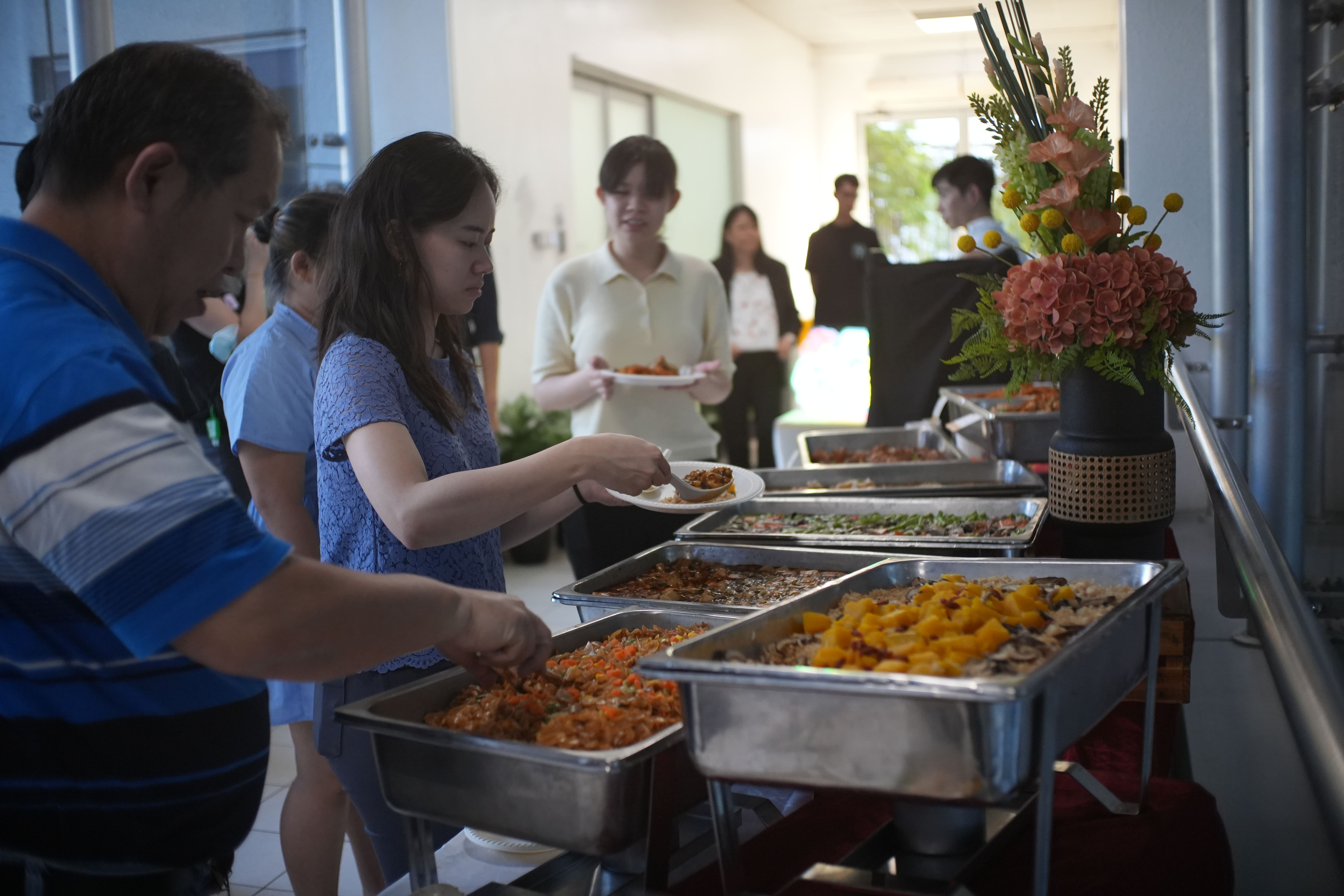 People serving themselves food from buffet trays at an indoor event, with a floral arrangement on the table.
