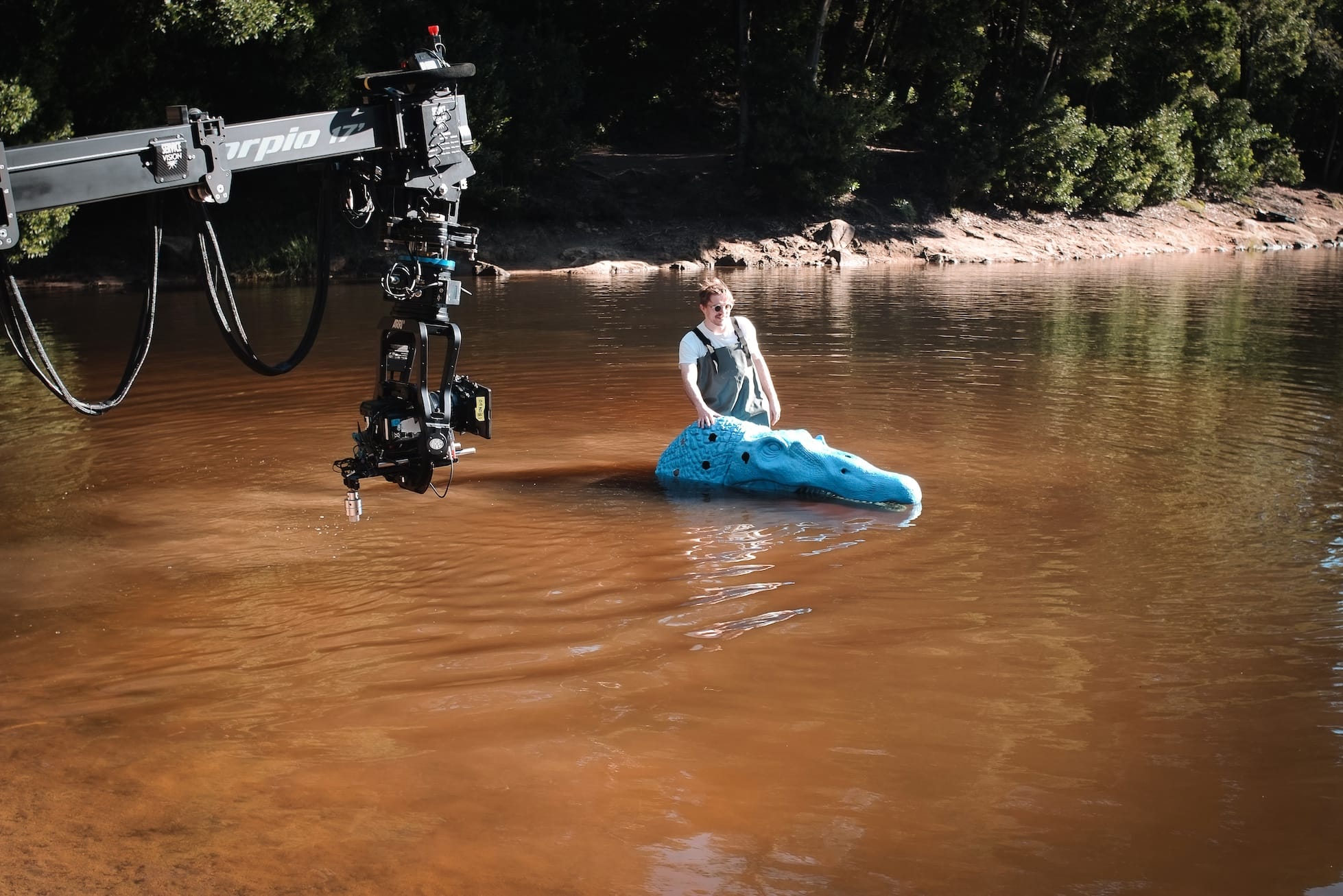 Person standing in a murky river beside a blue crocodile prop, filmed by a crane-mounted camera.