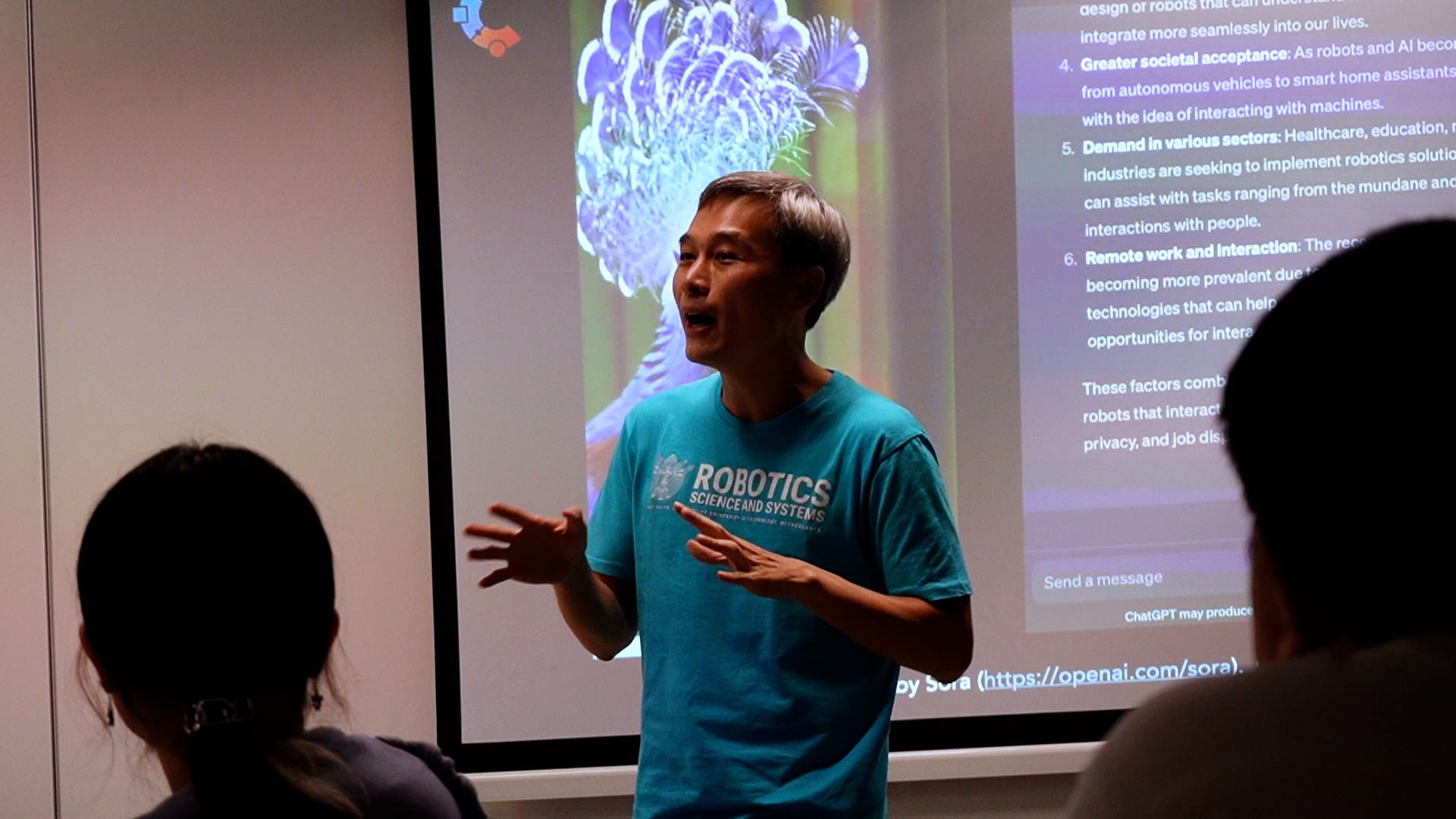 Man in a teal Robotics Science and Systems shirt speaks in front of a projected slide about robotics and AI.