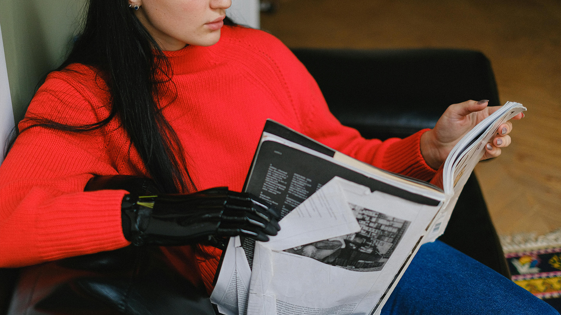 Young woman in a red sweater reading a magazine, holding it with one natural hand and one black prosthetic hand.