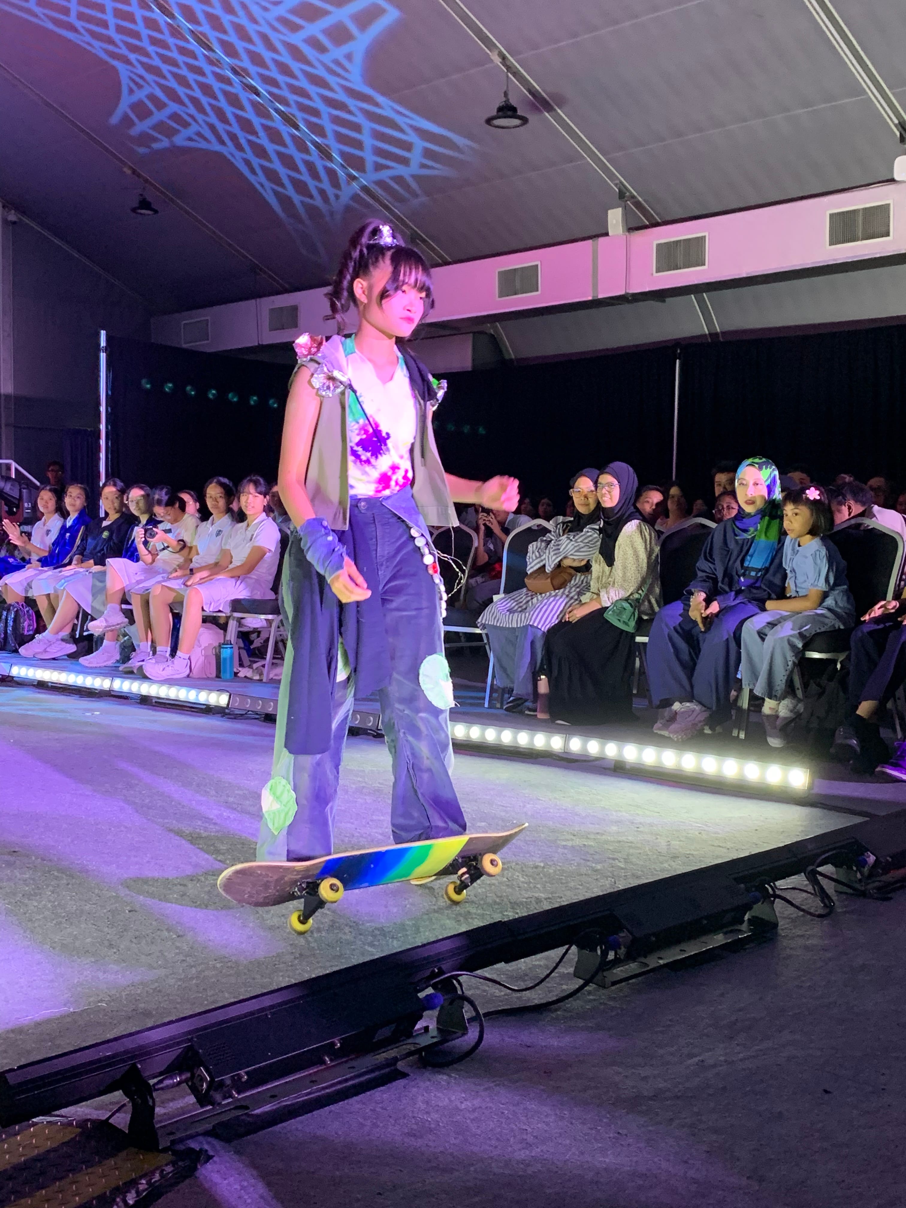 A young model rides a rainbow skateboard down a lit runway in purple flared jeans and floral top before a seated audience.