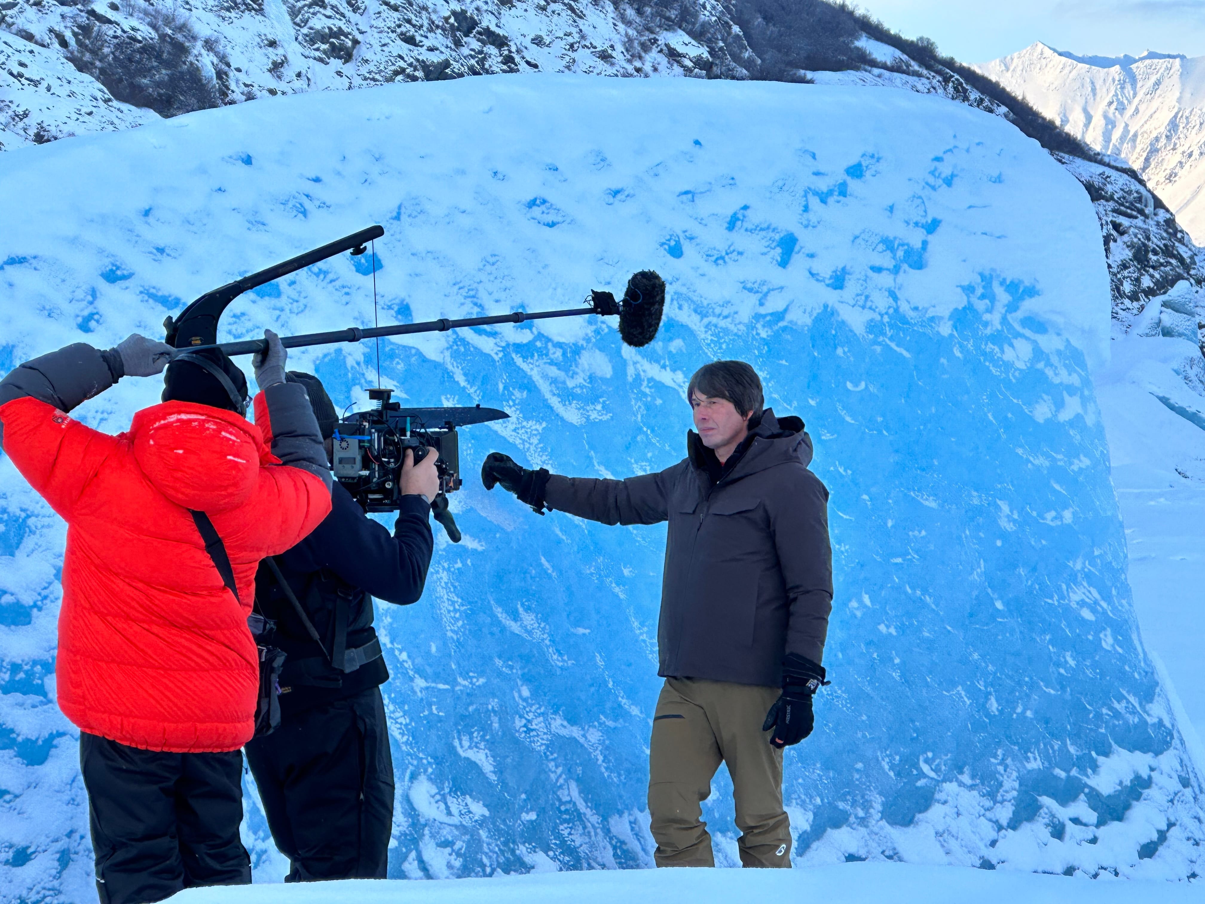 A film crew with a camera and boom mic records a man in a brown jacket in front of a large ice block outdoors.
