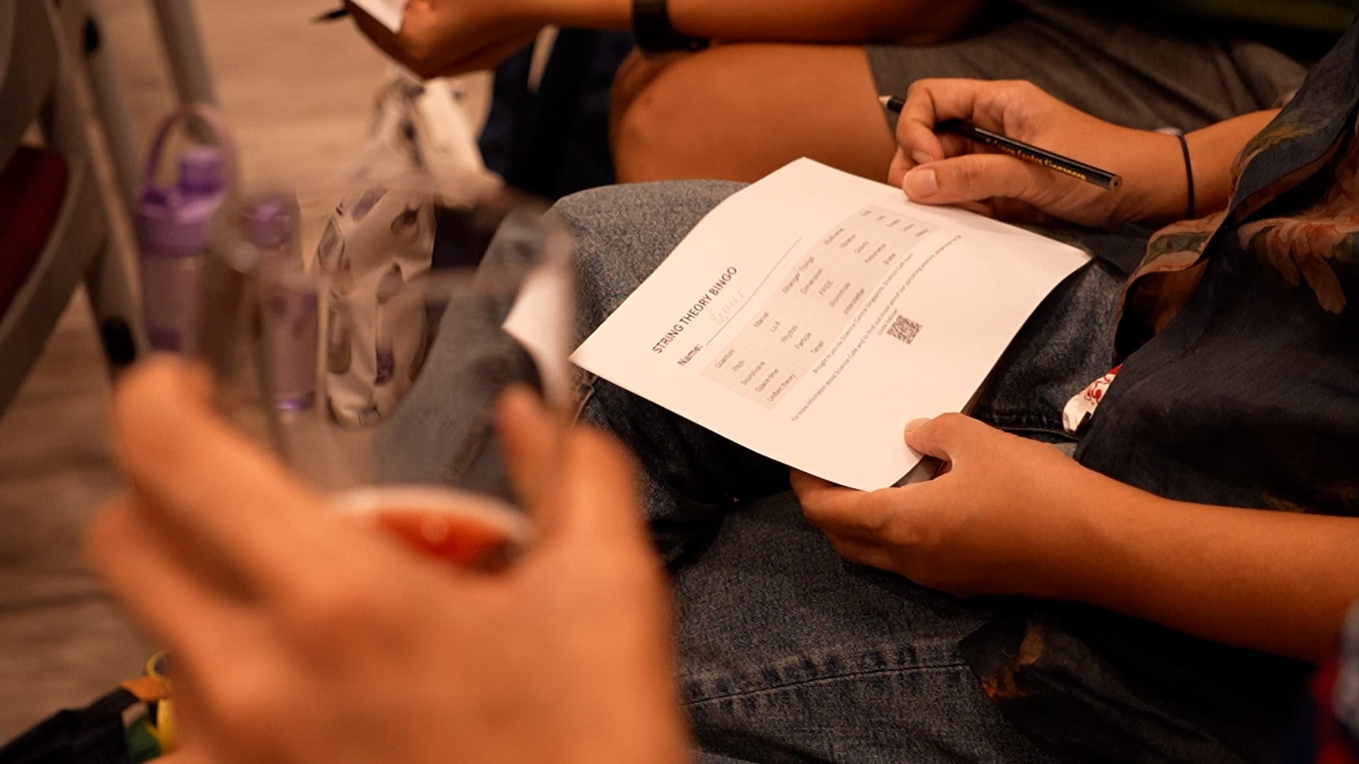 Person holding a printed "String Theory Bingo" sheet on their lap while another hand holds a pen nearby.