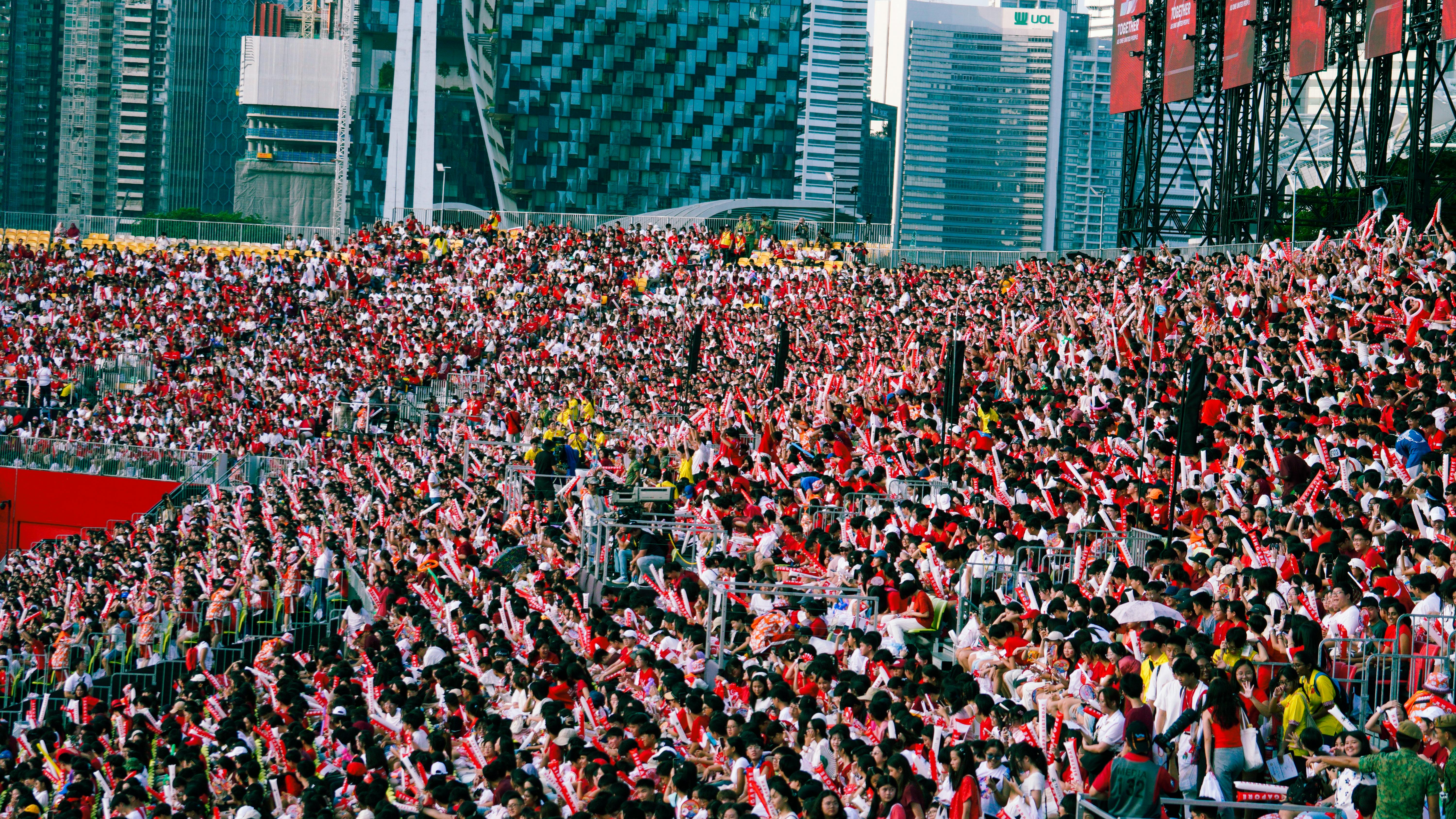 A massive crowd wearing red and white waves flags at an outdoor event with tall city buildings in the background.