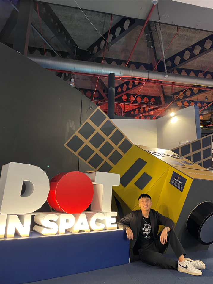 Jarrod Chua sitting beside a large "DOT IN SPACE" sign with a yellow satellite model at an indoor space-themed exhibit.