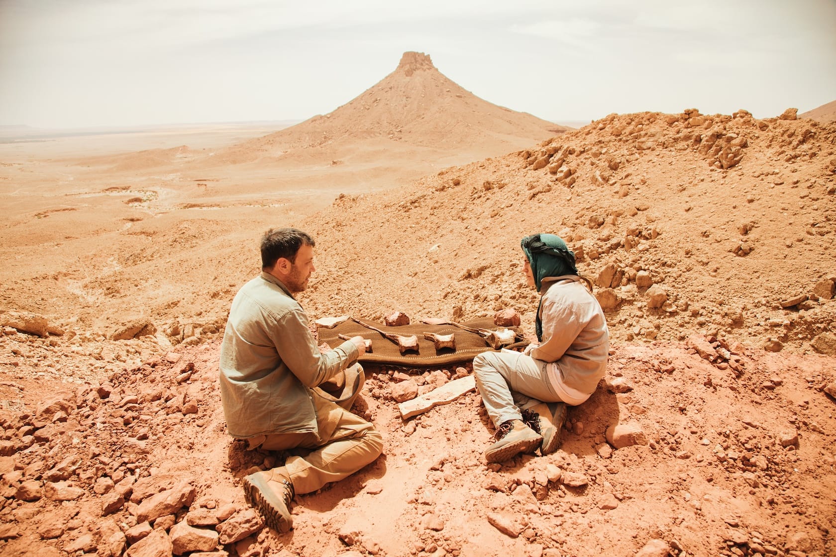 Two people sit on rocky desert ground examining bone fragments on a cloth, with a pointed mesa in the background.