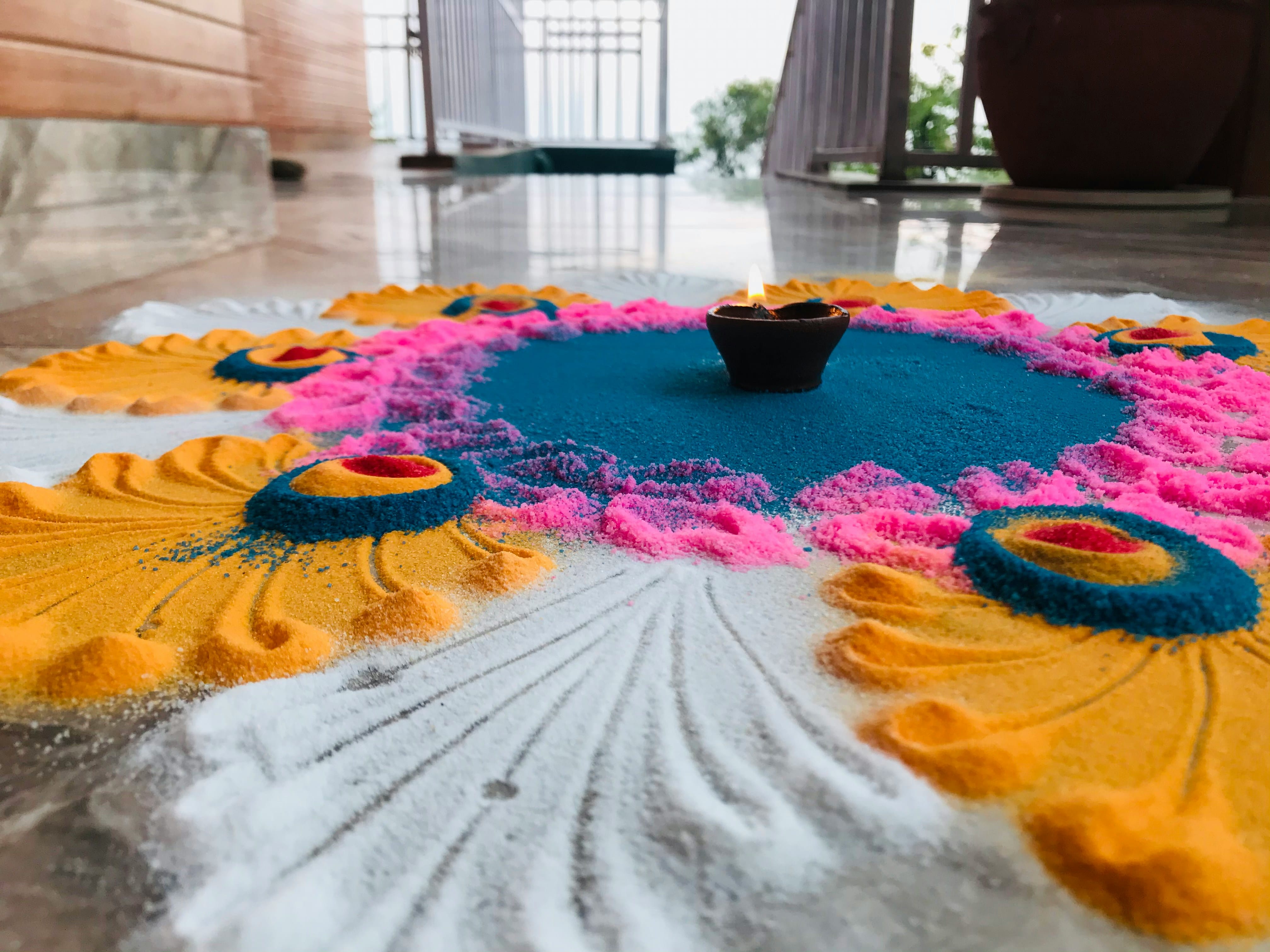 Colorful rangoli made of yellow, pink, blue, and white powder on a floor with a lit clay diya in the center.