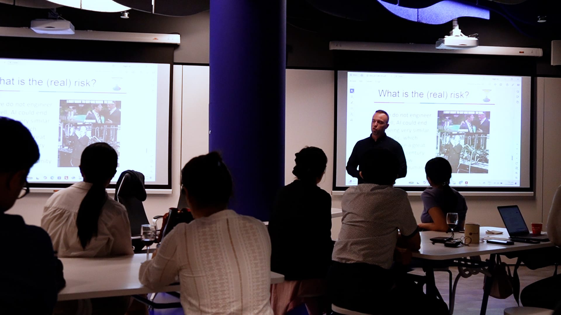 A man presents to seated attendees in a room with two screens displaying "What is the (real) risk?"