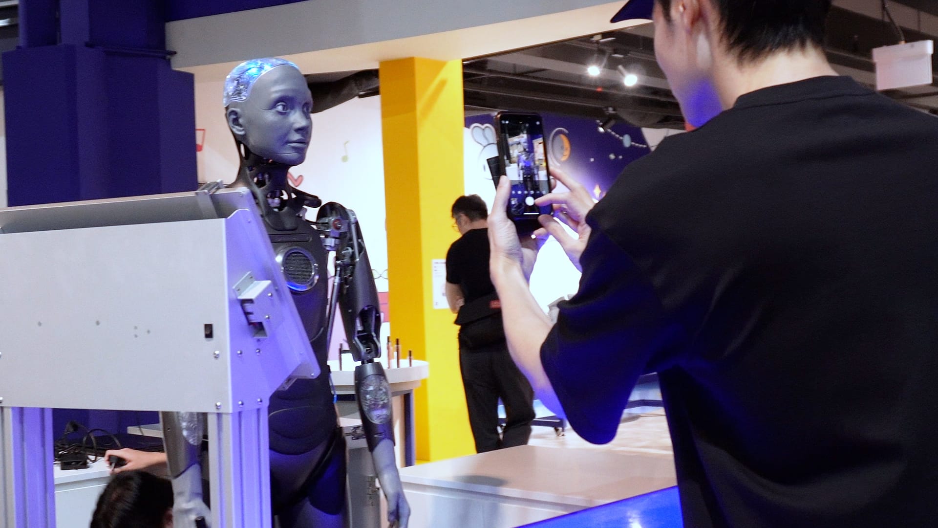 A person photographs a humanoid robot with a realistic face at an indoor tech exhibition.