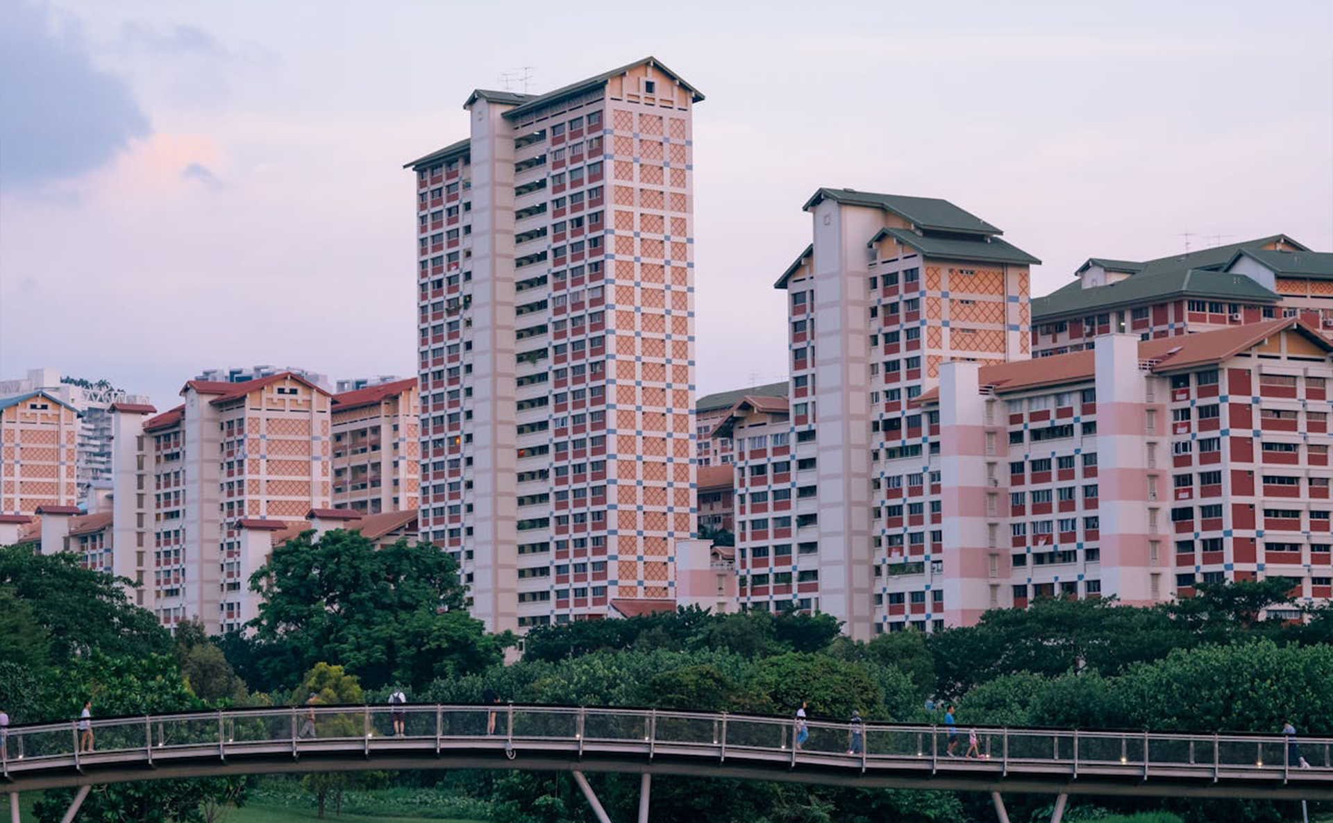 Tall red and beige residential tower blocks behind green trees, with a curved pedestrian bridge in the foreground at dusk.