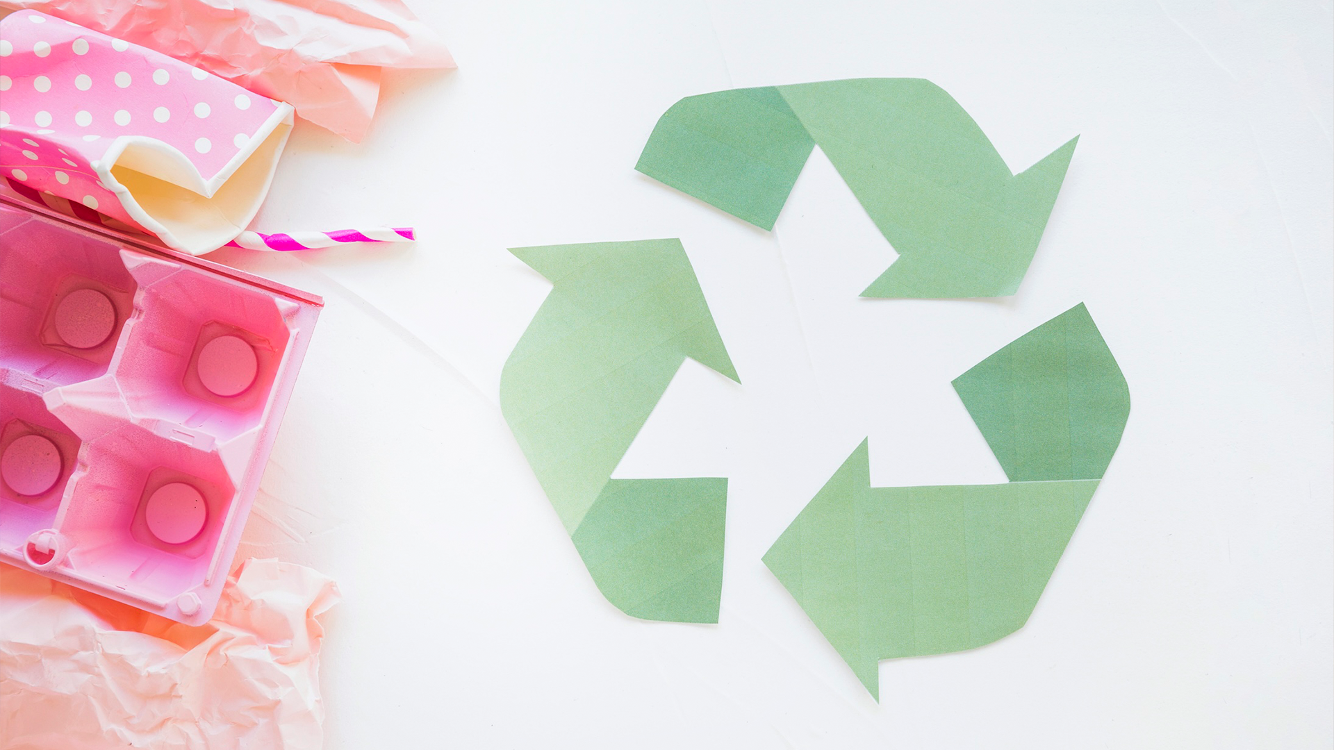 Green paper recycling symbol on white background beside pink polka-dot cup, straw, and egg carton.