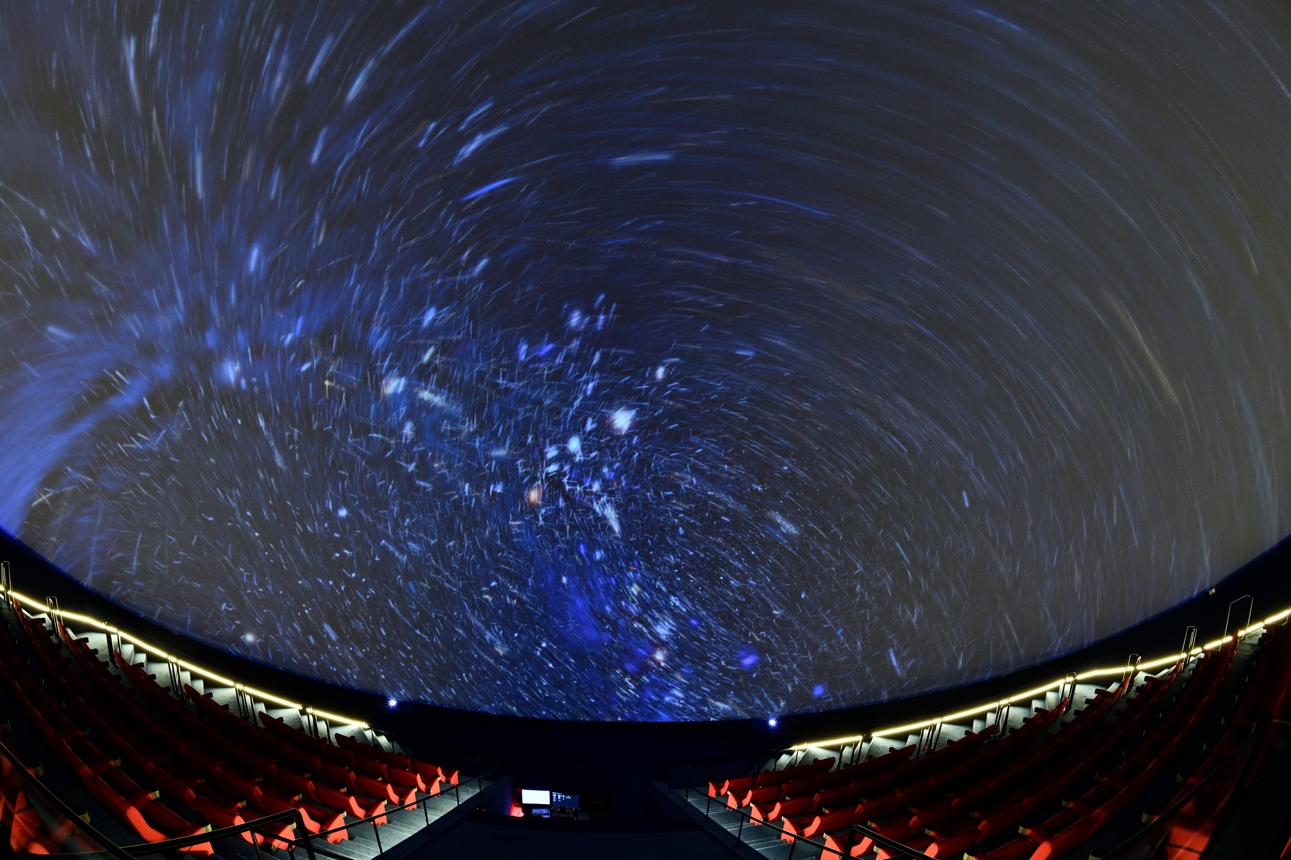 Planetarium dome with swirling blue star trails projected on ceiling above rows of red seats.