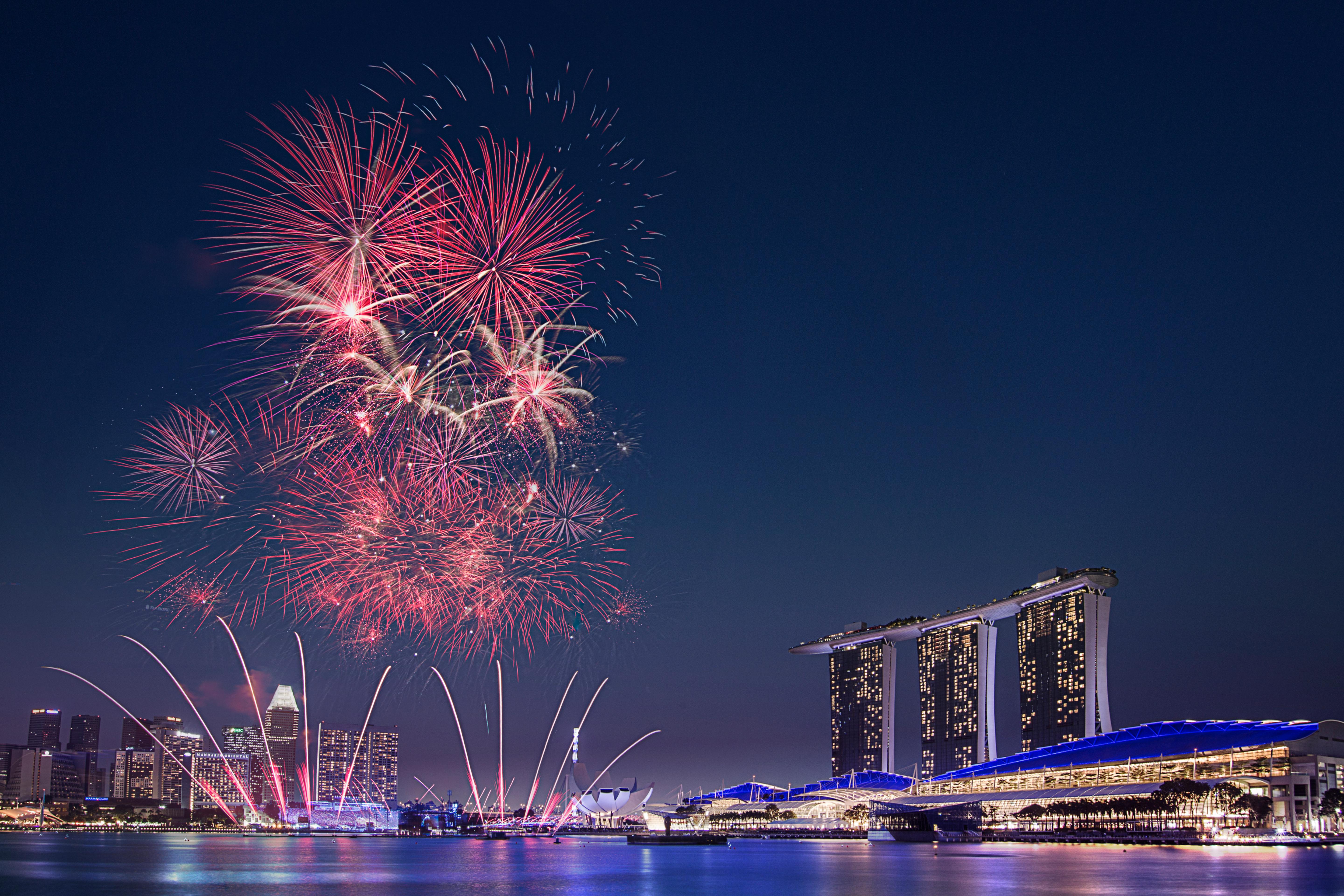 Red fireworks burst over Singapore's Marina Bay Sands hotel and illuminated skyline reflected in the water at night.