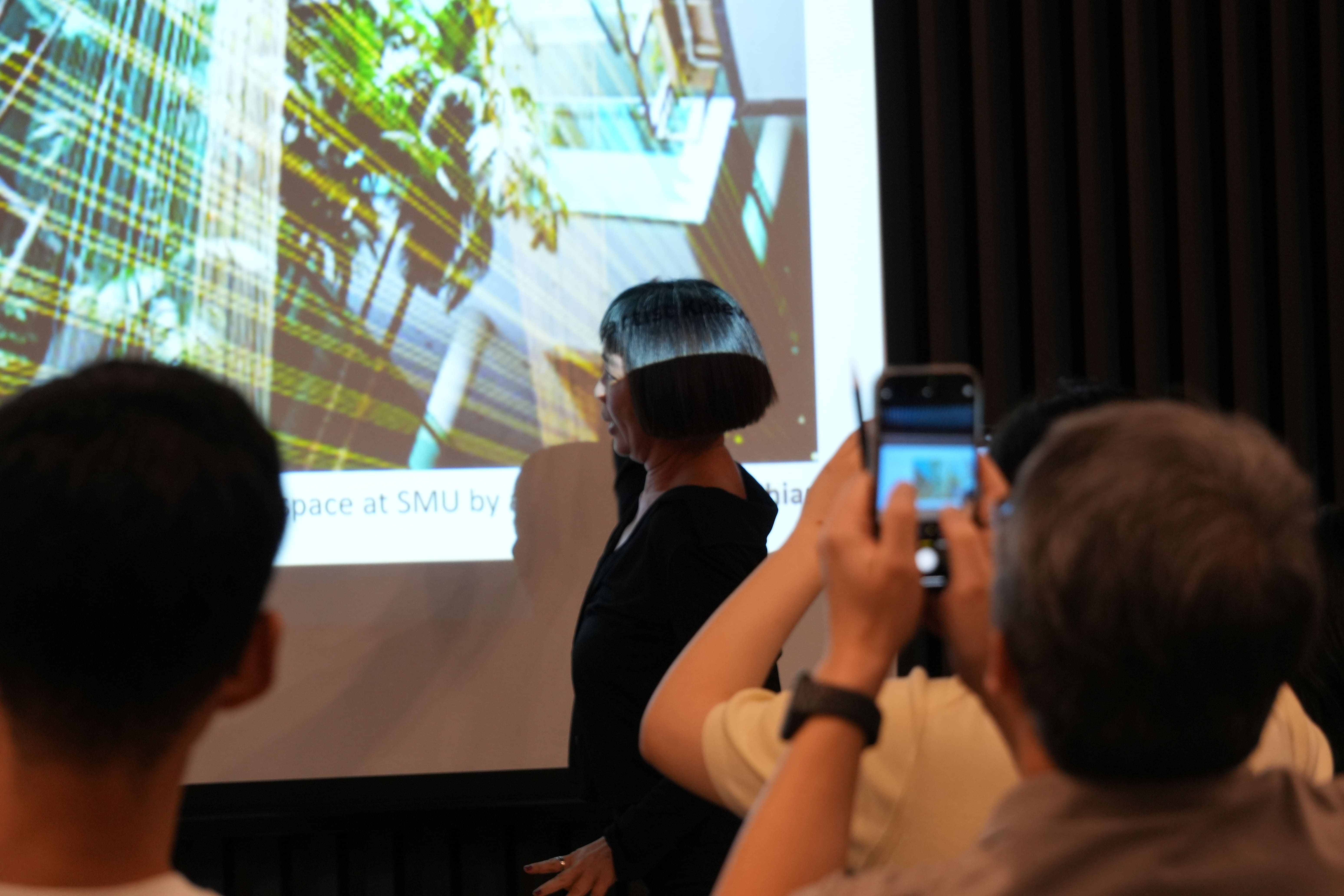 A woman in black presents in front of a projected slide showing an architectural space at SMU, as an audience member photographs with a phone.