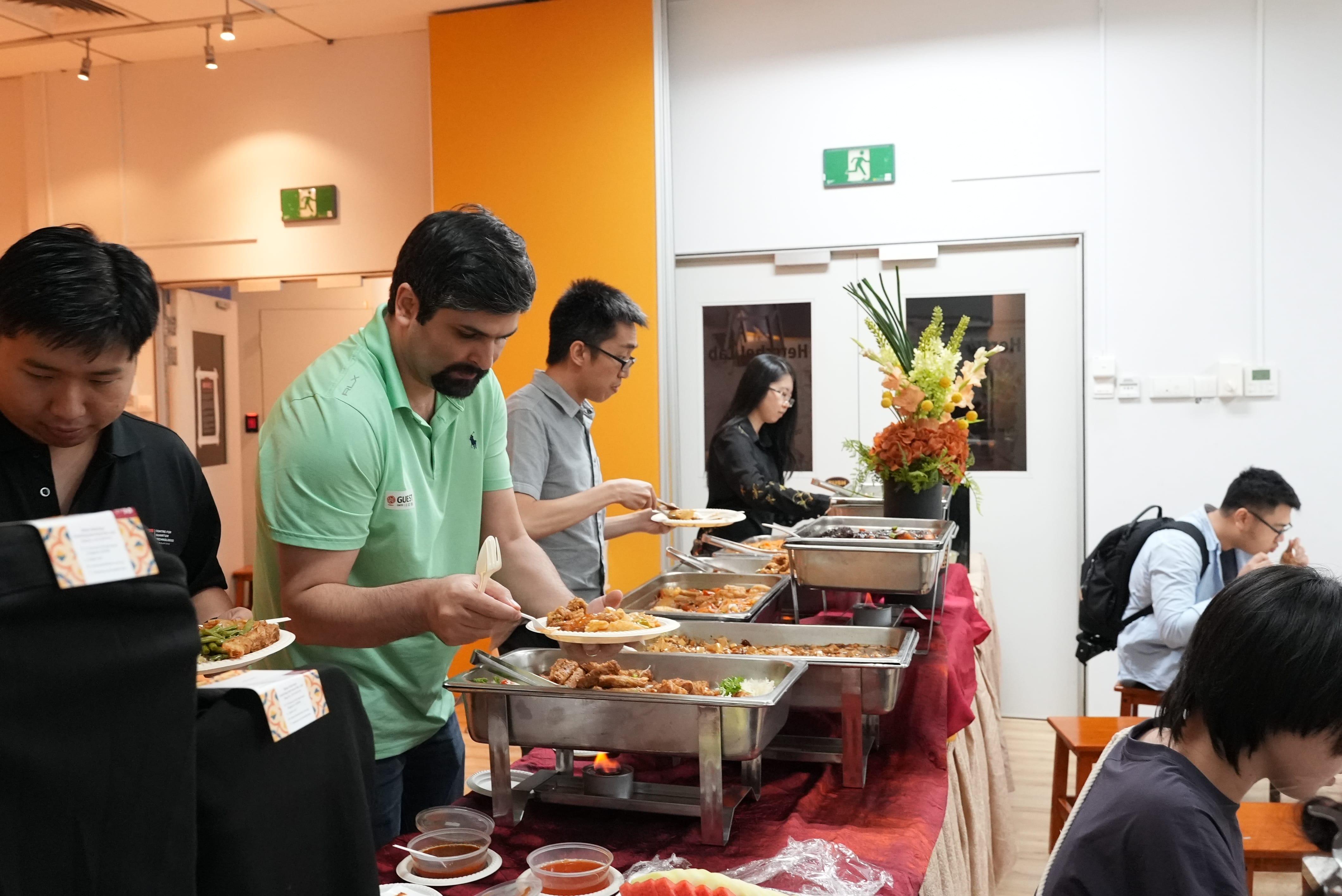 People serving themselves food from chafing dishes at an indoor buffet with a floral centerpiece and orange walls.