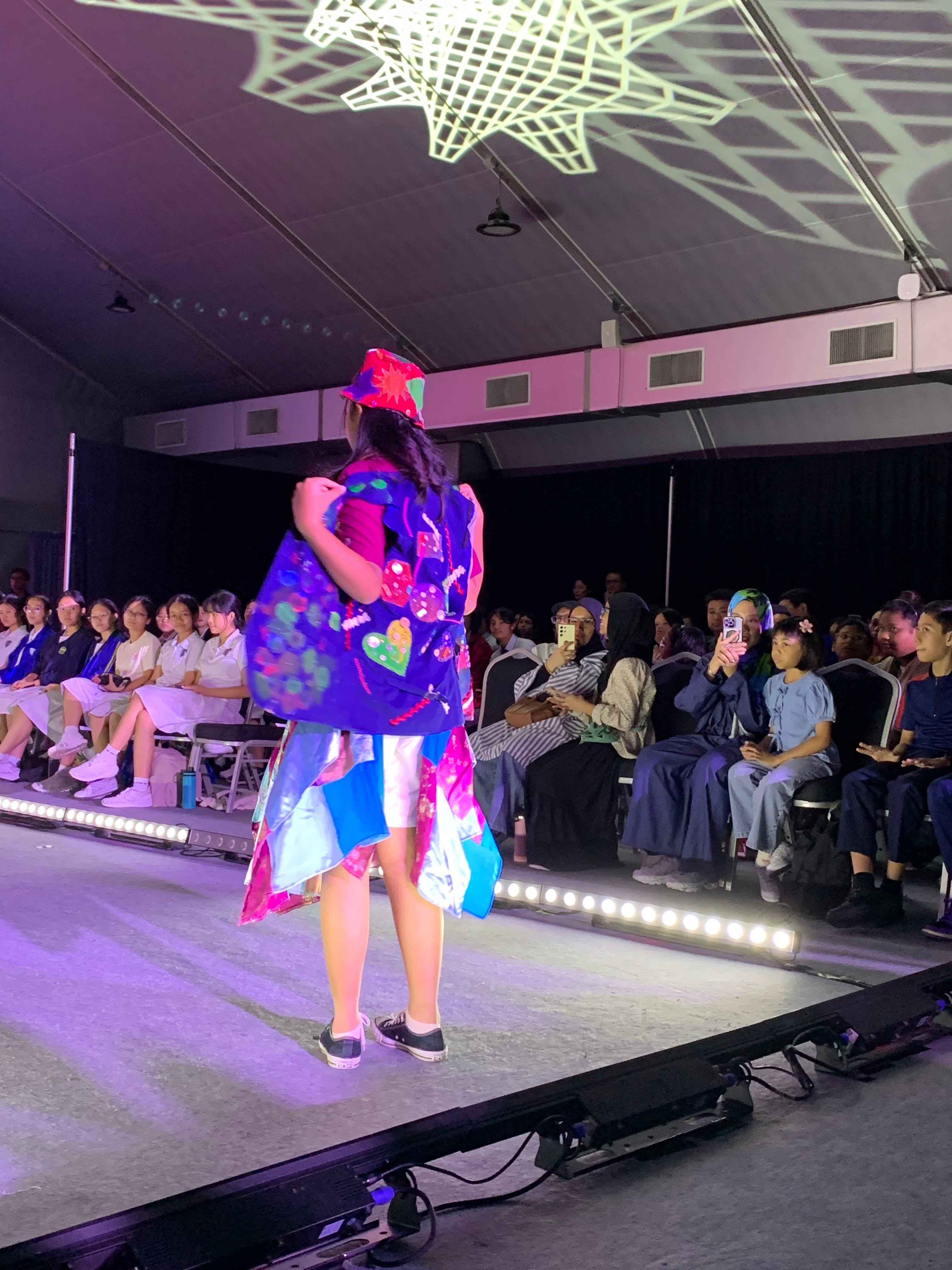 A young model walks a lit runway in a colorful painted outfit and red bucket hat, with a seated audience watching.