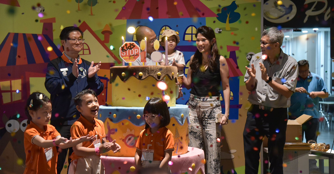 Children in orange shirts and adults celebrate around a large decorated cake with candles, confetti falling, colorful mural behind them.