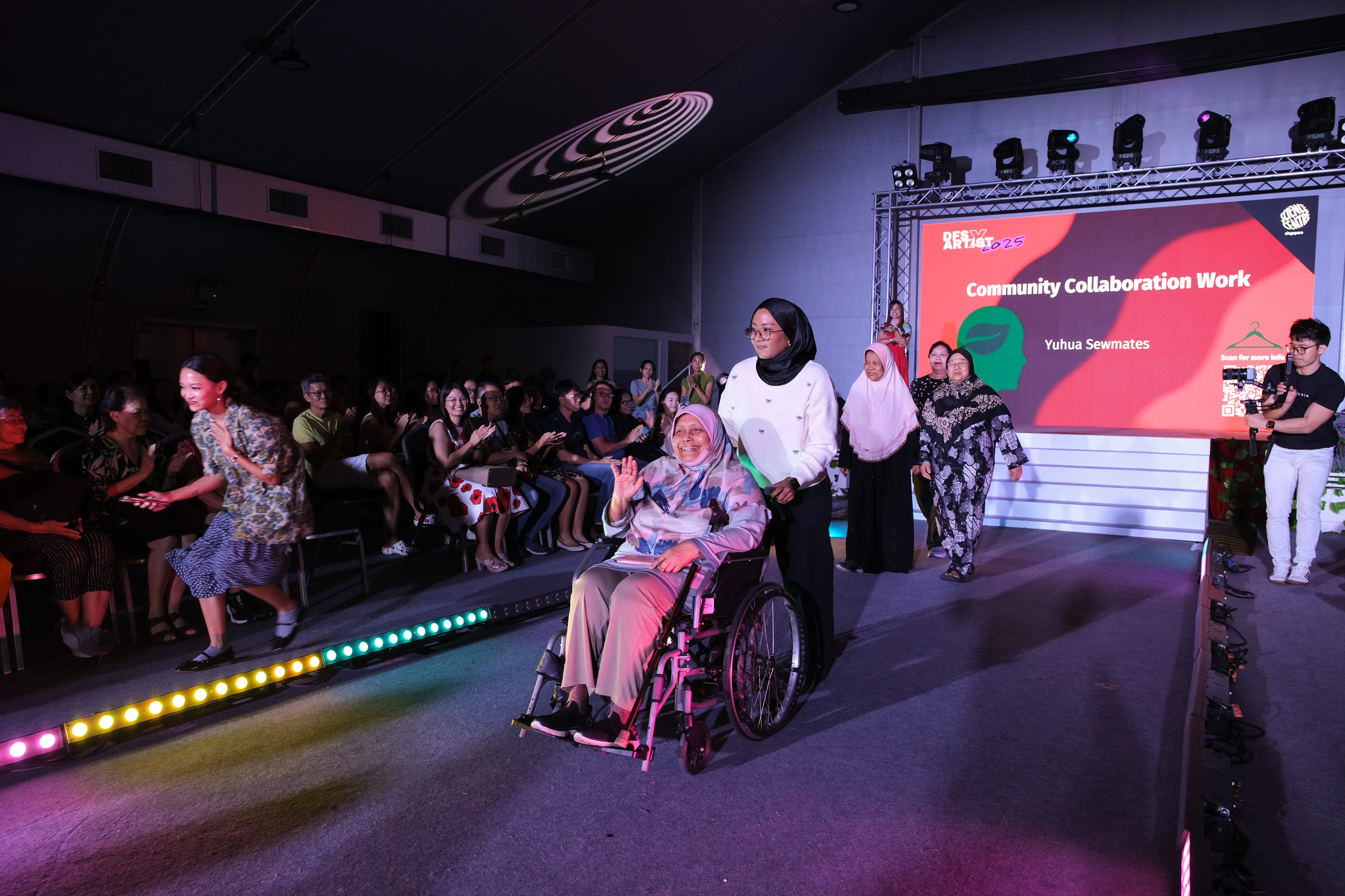 A woman in a wheelchair waves on a runway at a community event, pushed by another woman, with an audience applauding.