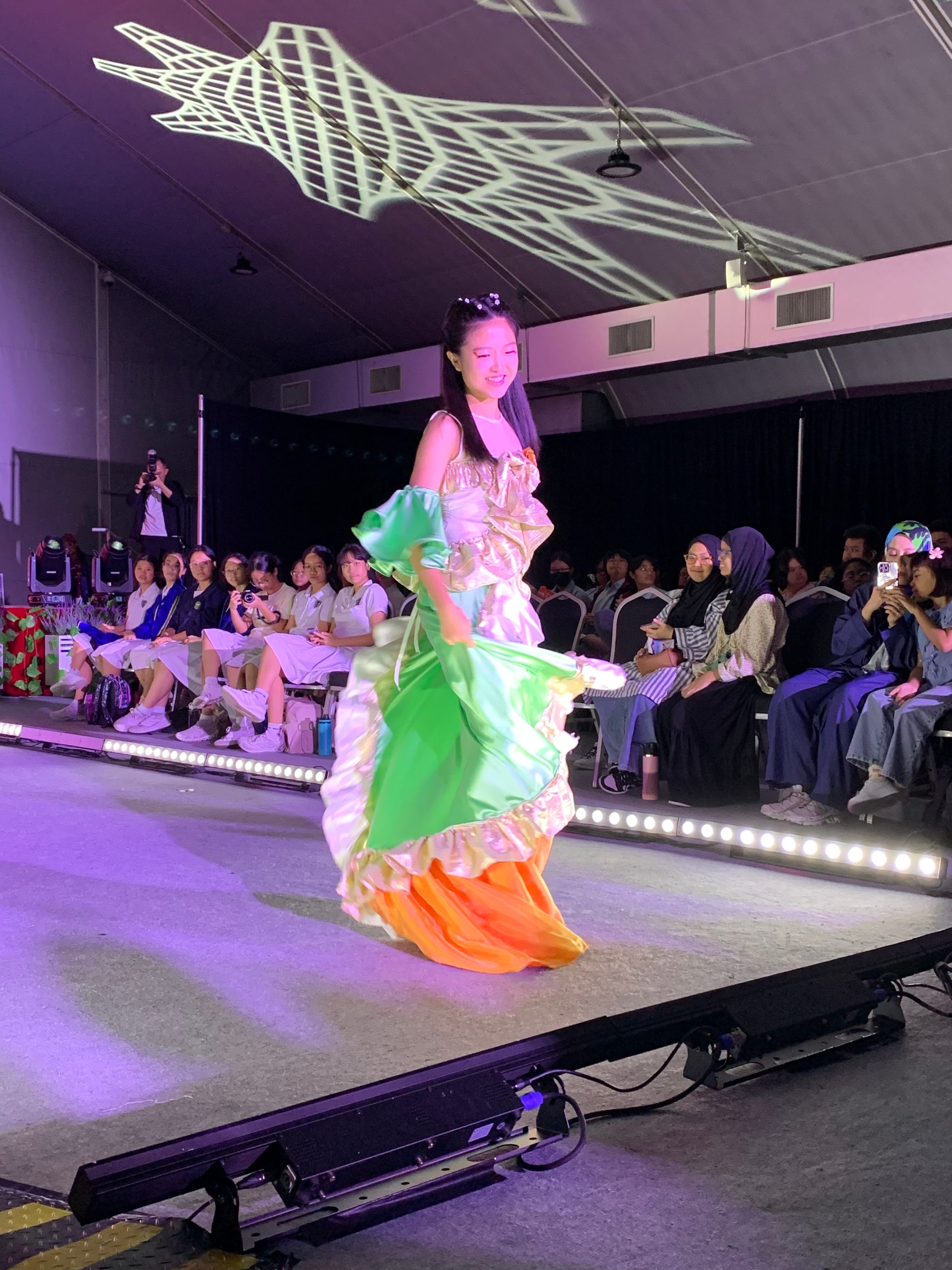 A young woman in a green and orange ruffled gown walks a lit runway while seated spectators watch and photograph.