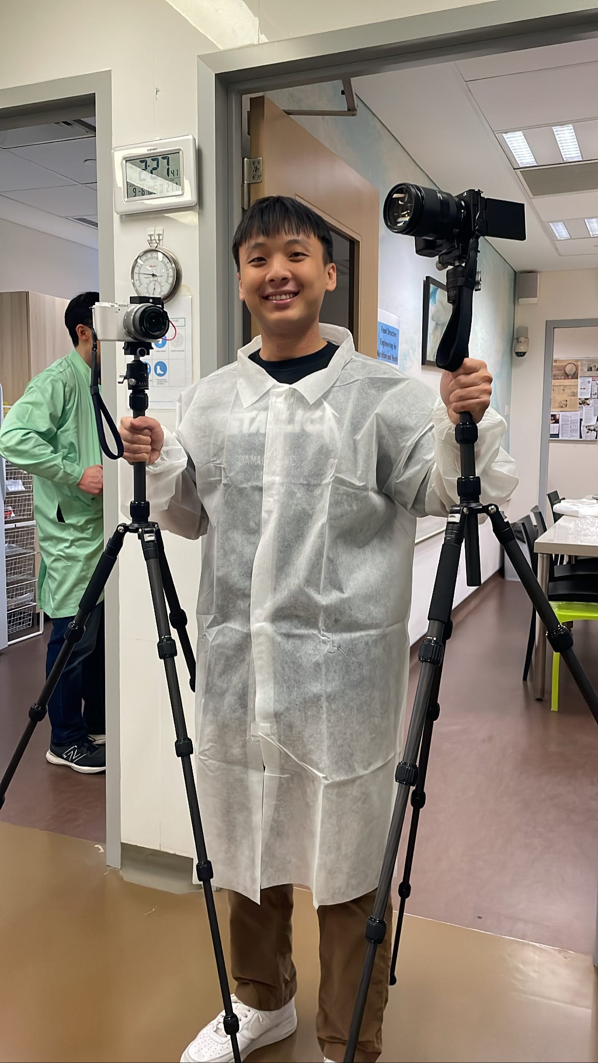Jarrod Chua holds two camera tripods with cameras in an indoor setting.