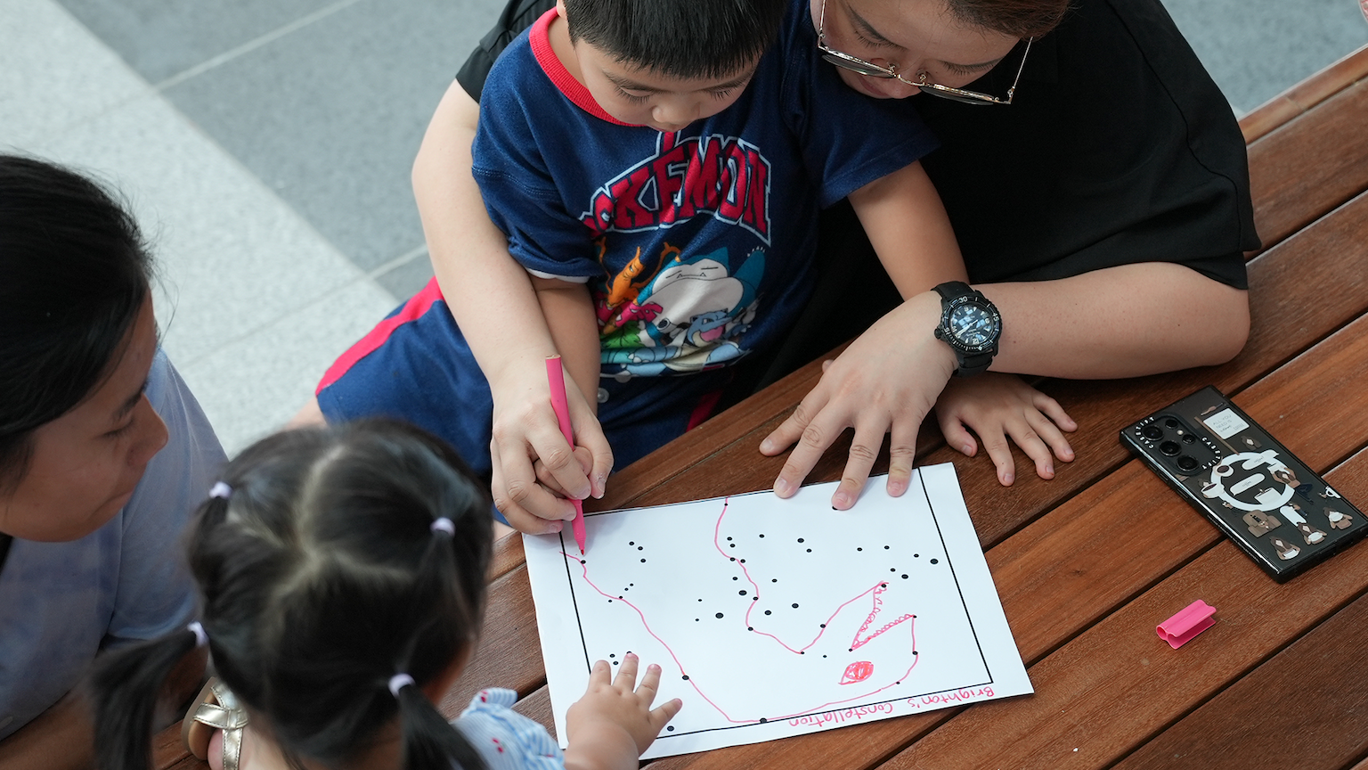 A boy draws pink lines connecting dots on a constellation worksheet at a wooden table with an adult and young girl watching.