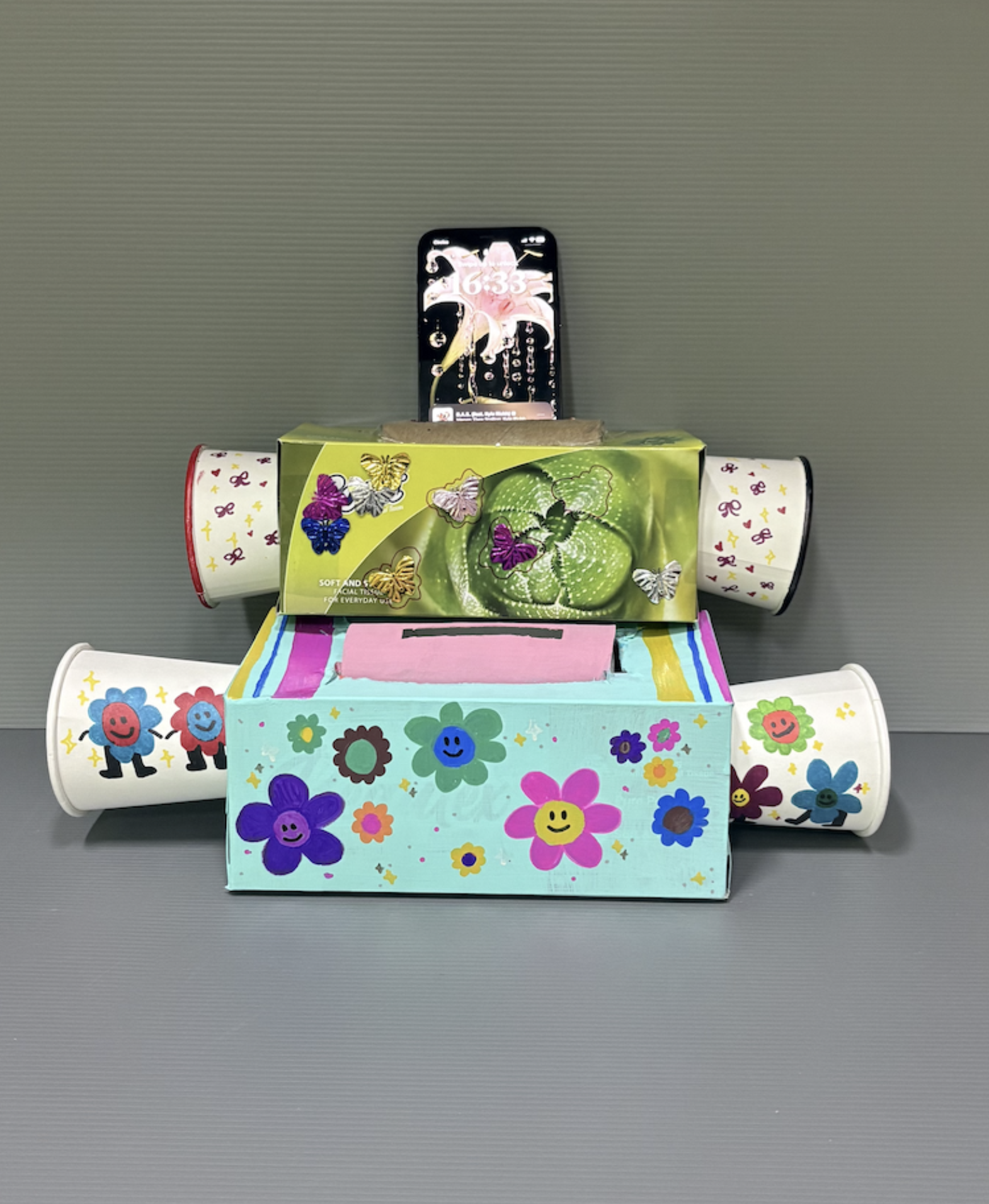 Two decorated tissue boxes with smiley flower designs, four cups, and a phone balanced on top forming a robot shape.