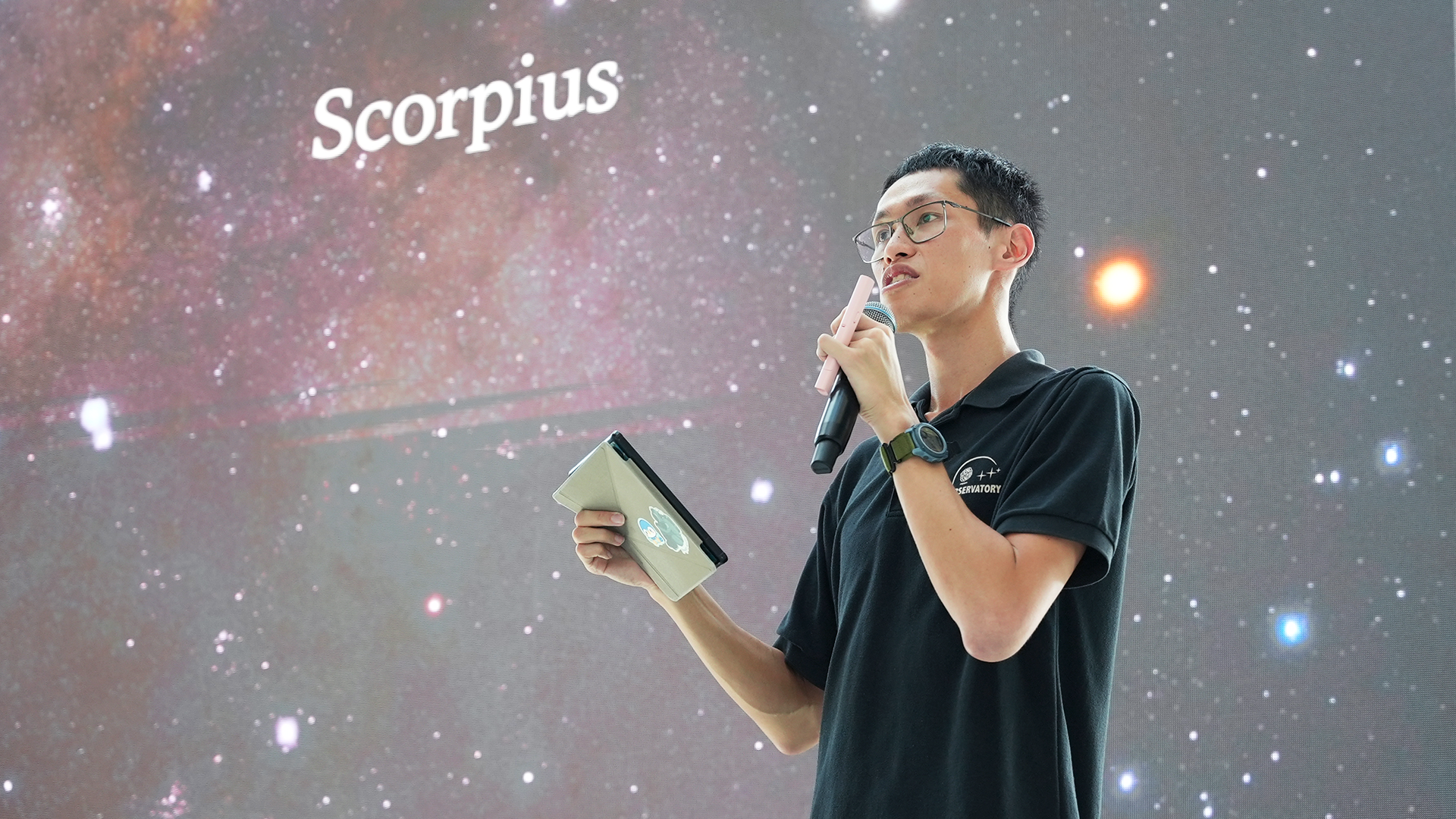 Meng holding a microphone and tablet presents in front of a starry projection screen showing the word Scorpius.