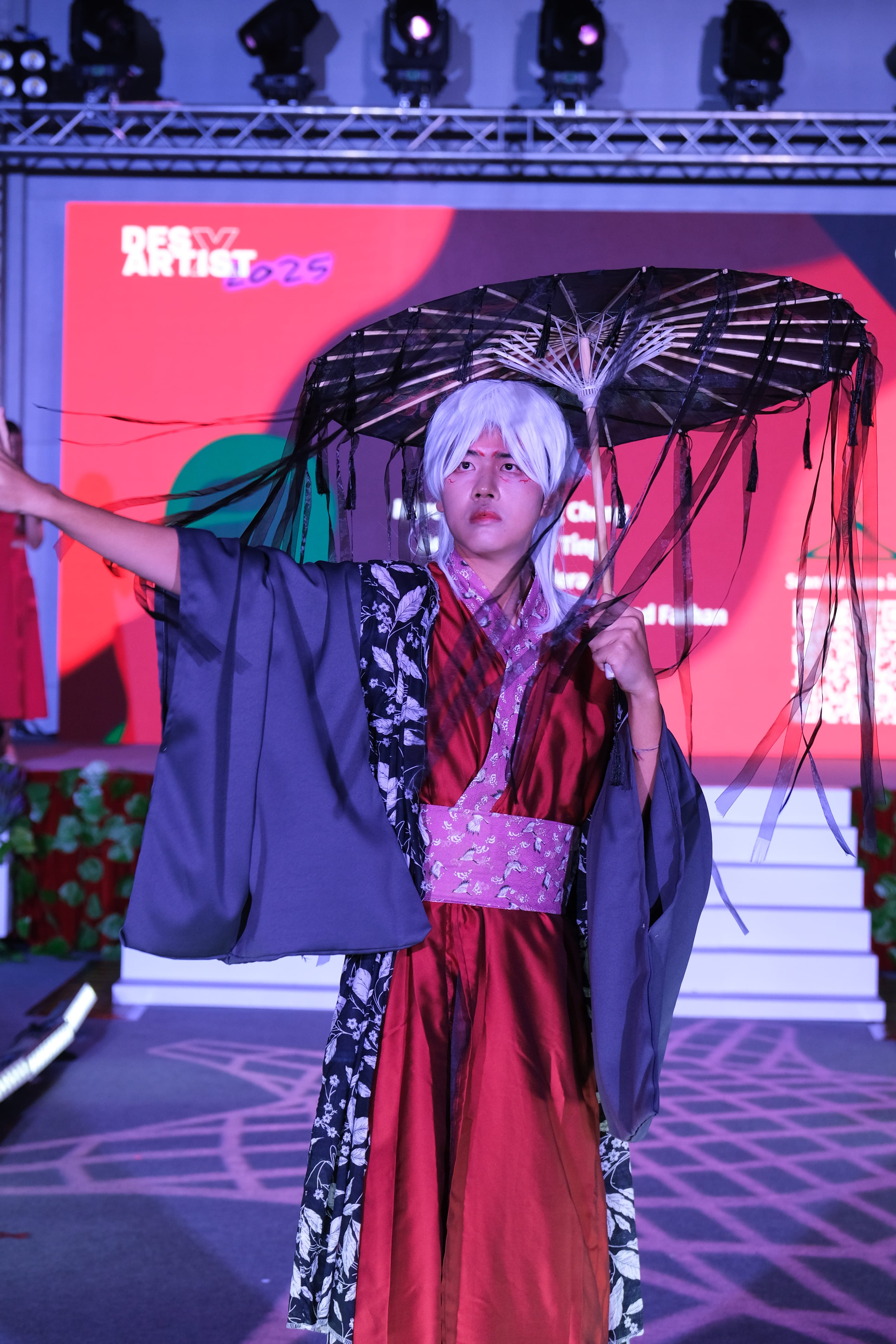 Person in white wig and red kimono holds a dark tattered parasol on a lit stage at a fashion event.