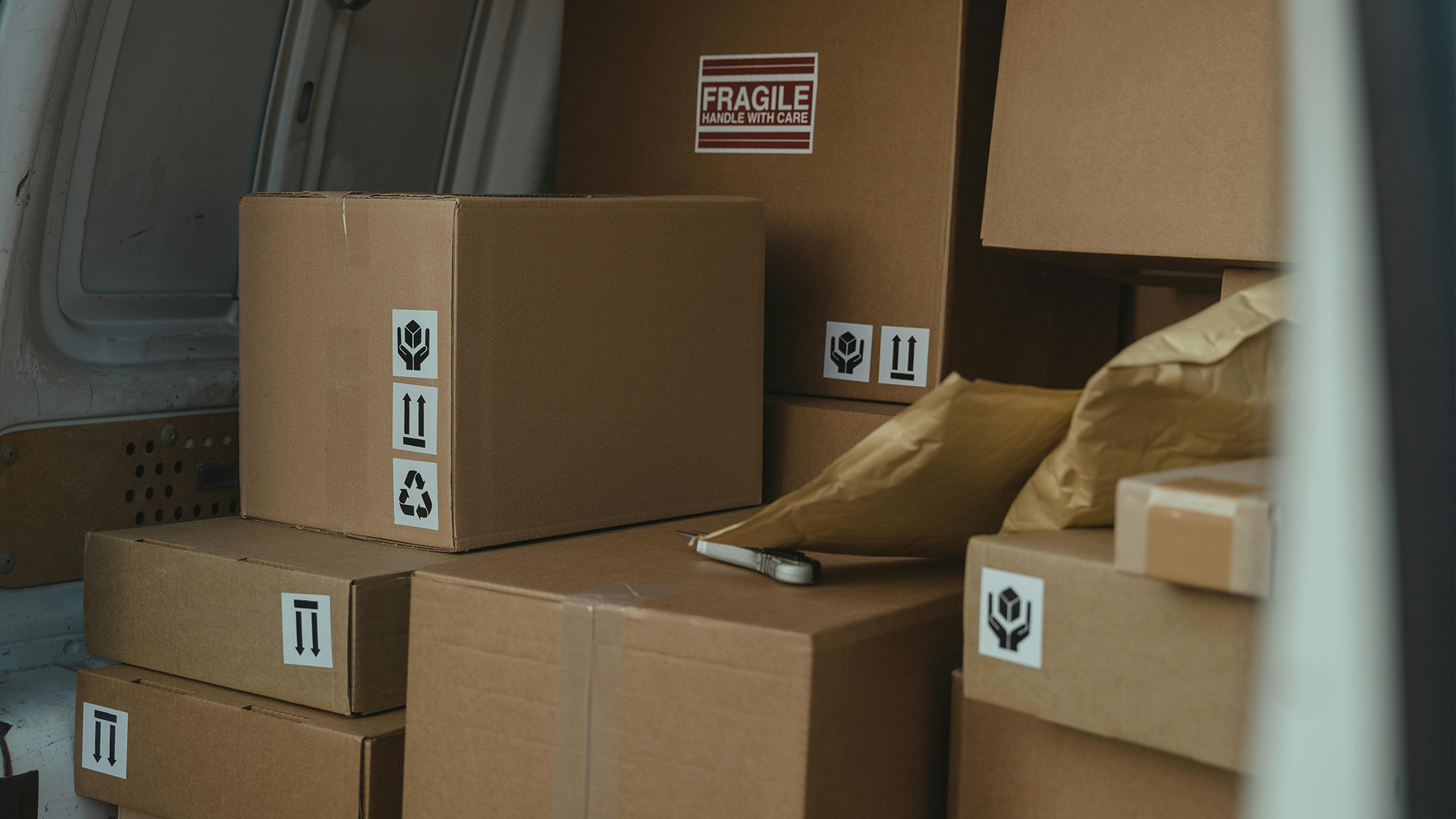 Cardboard boxes stacked inside a delivery van, one labeled Fragile Handle with Care, with padded envelopes nearby.