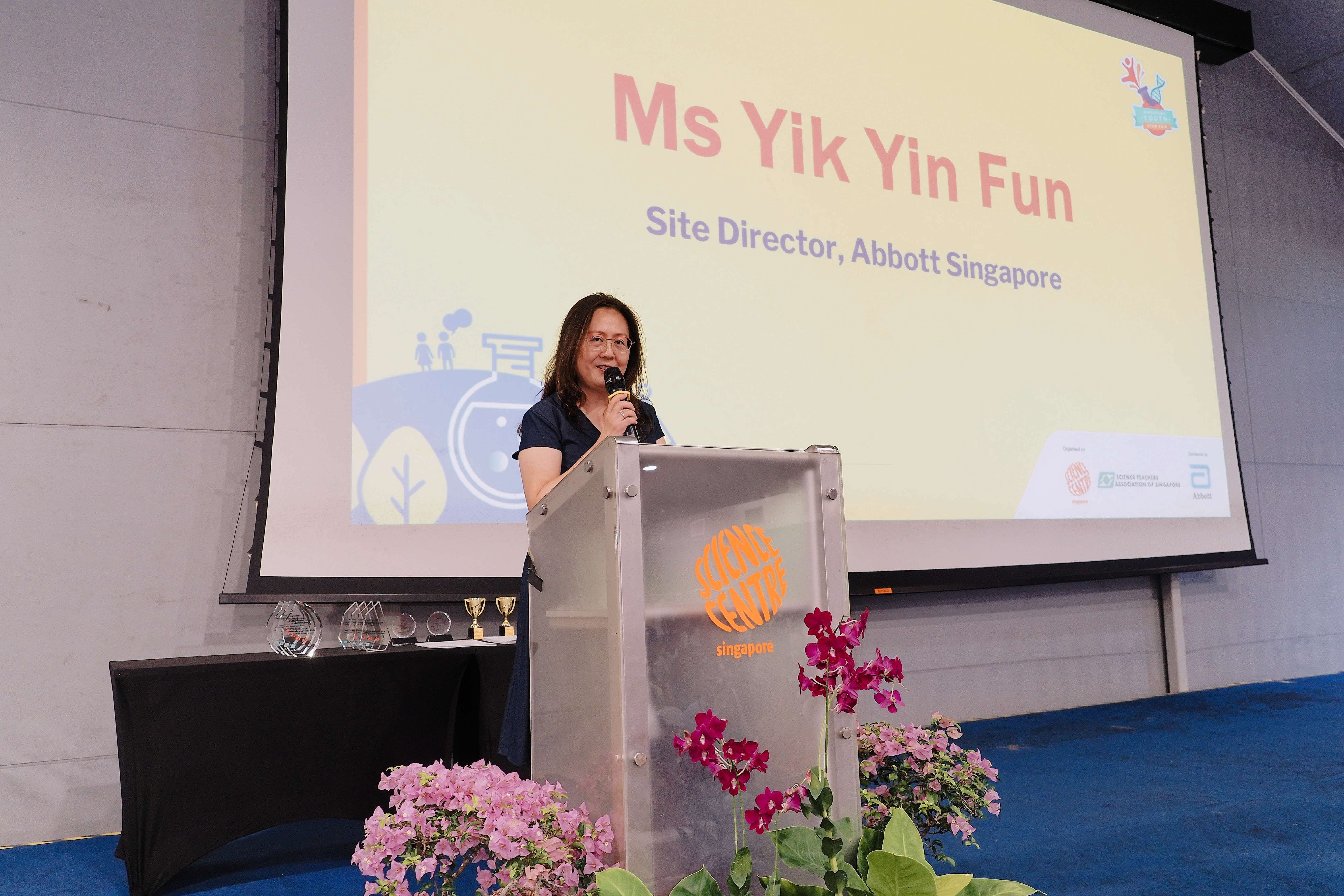 Woman speaking at a Science Centre Singapore podium, with a slide showing "Ms Yik Yin Fun, Site Director, Abbott Singapore."