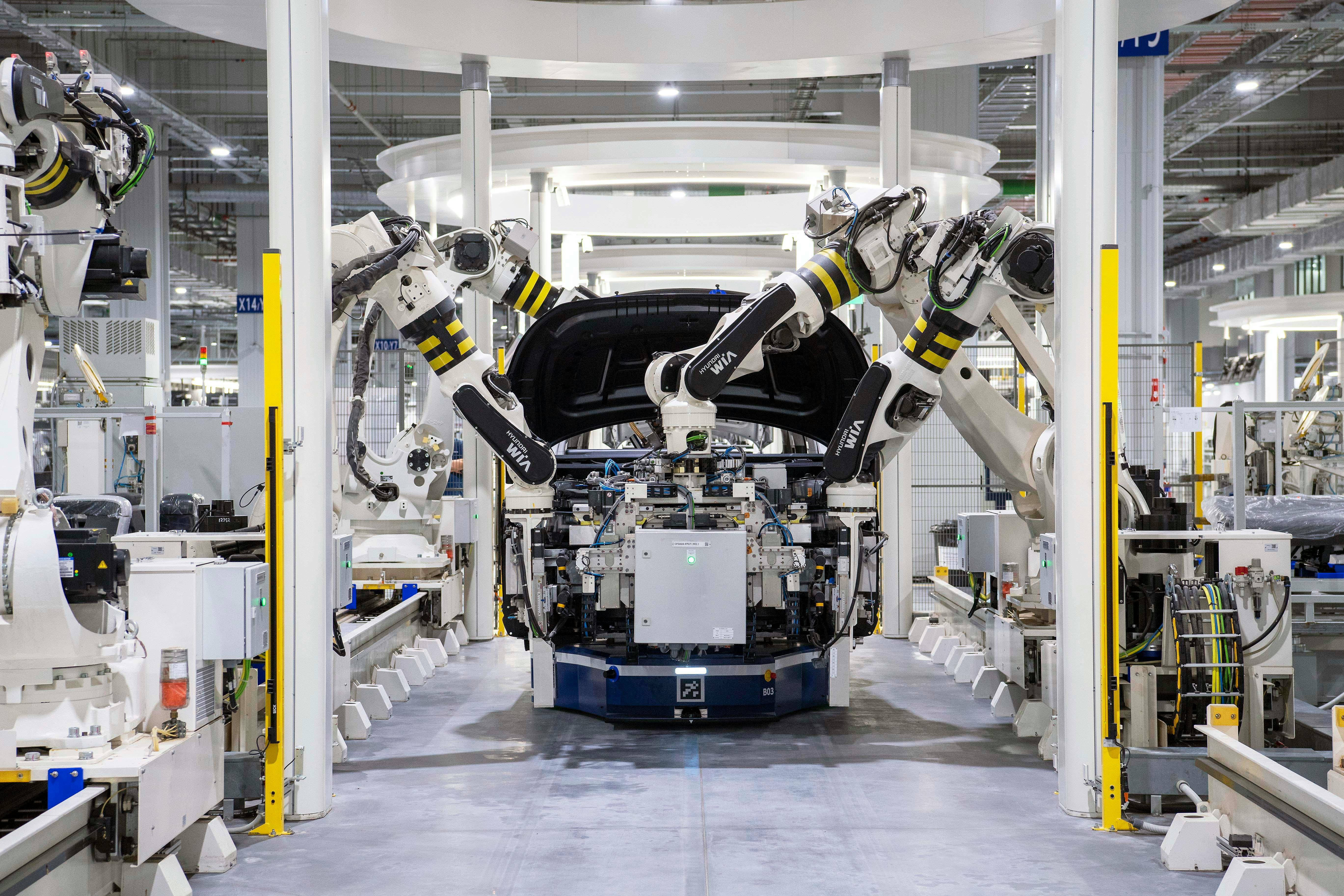 Large white robotic arms with yellow markings assembling a car body on an automated factory floor.
