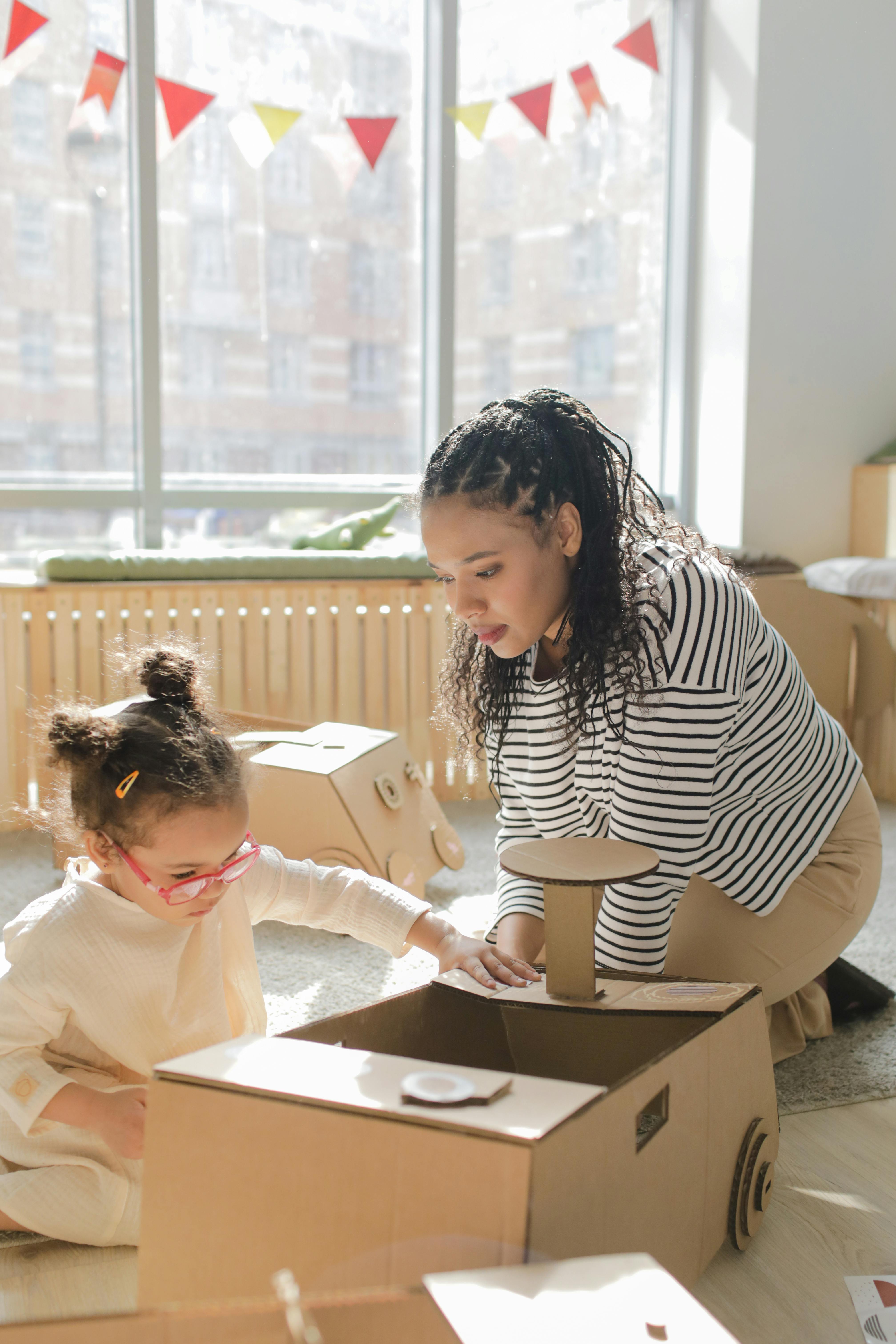 A woman and a young girl with pink glasses play together with a cardboard toy car on a sunny room floor.