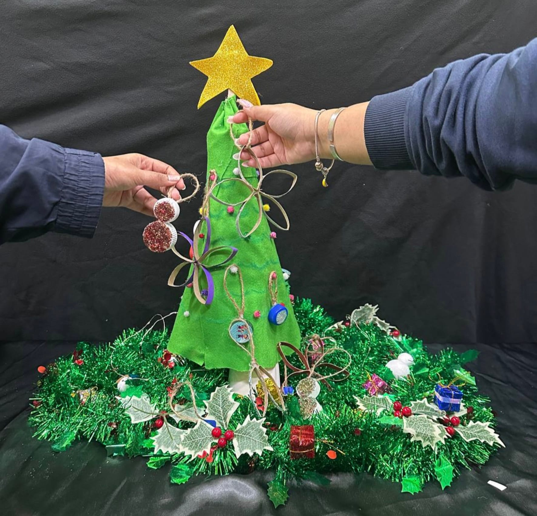 Two people decorating a handmade green fabric Christmas tree with a gold star, ornaments, and a holly wreath base.