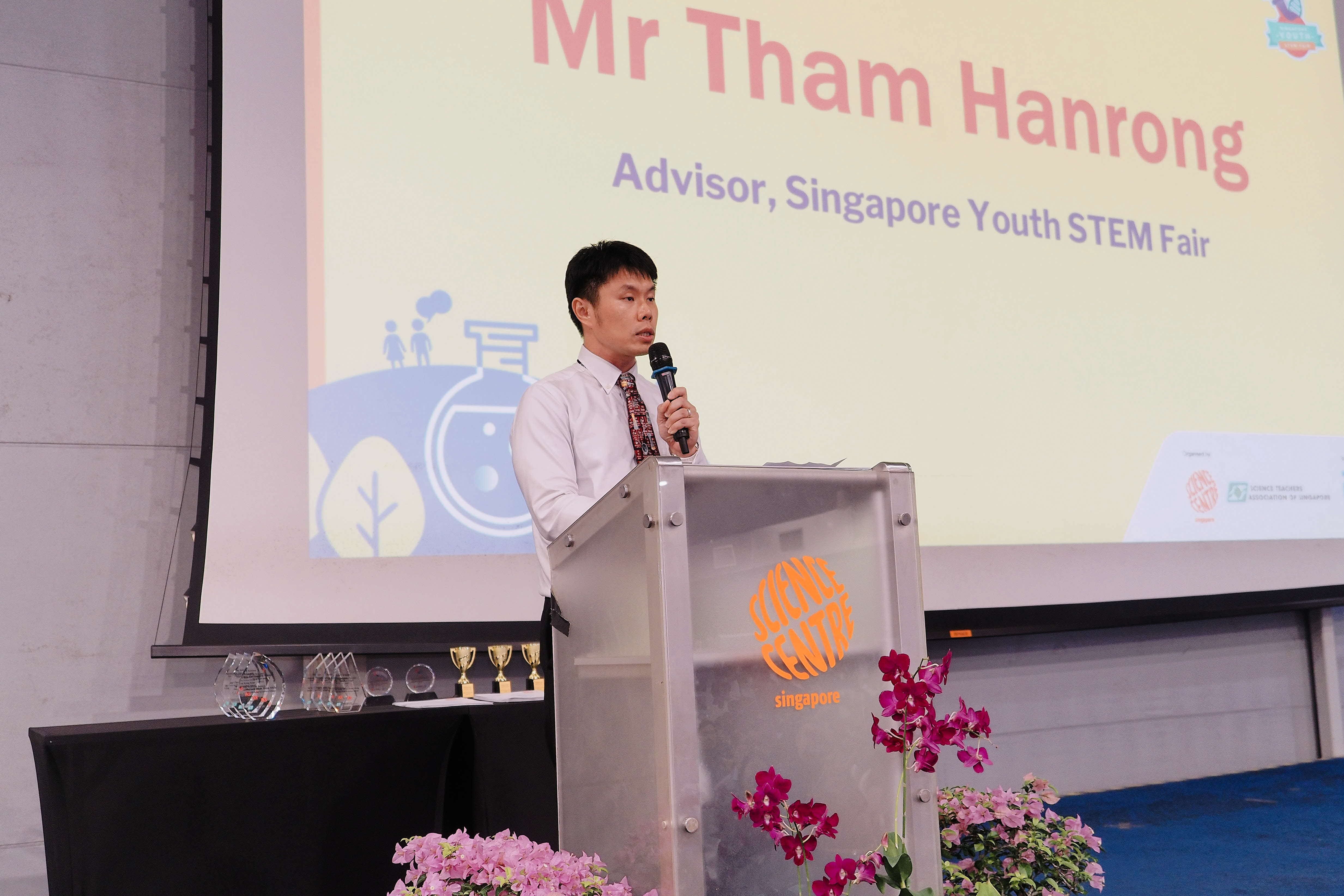 Man in white shirt speaking at a Science Centre Singapore podium with a slide reading "Mr Tham Hanrong, Advisor, Singapore Youth STEM Fair."