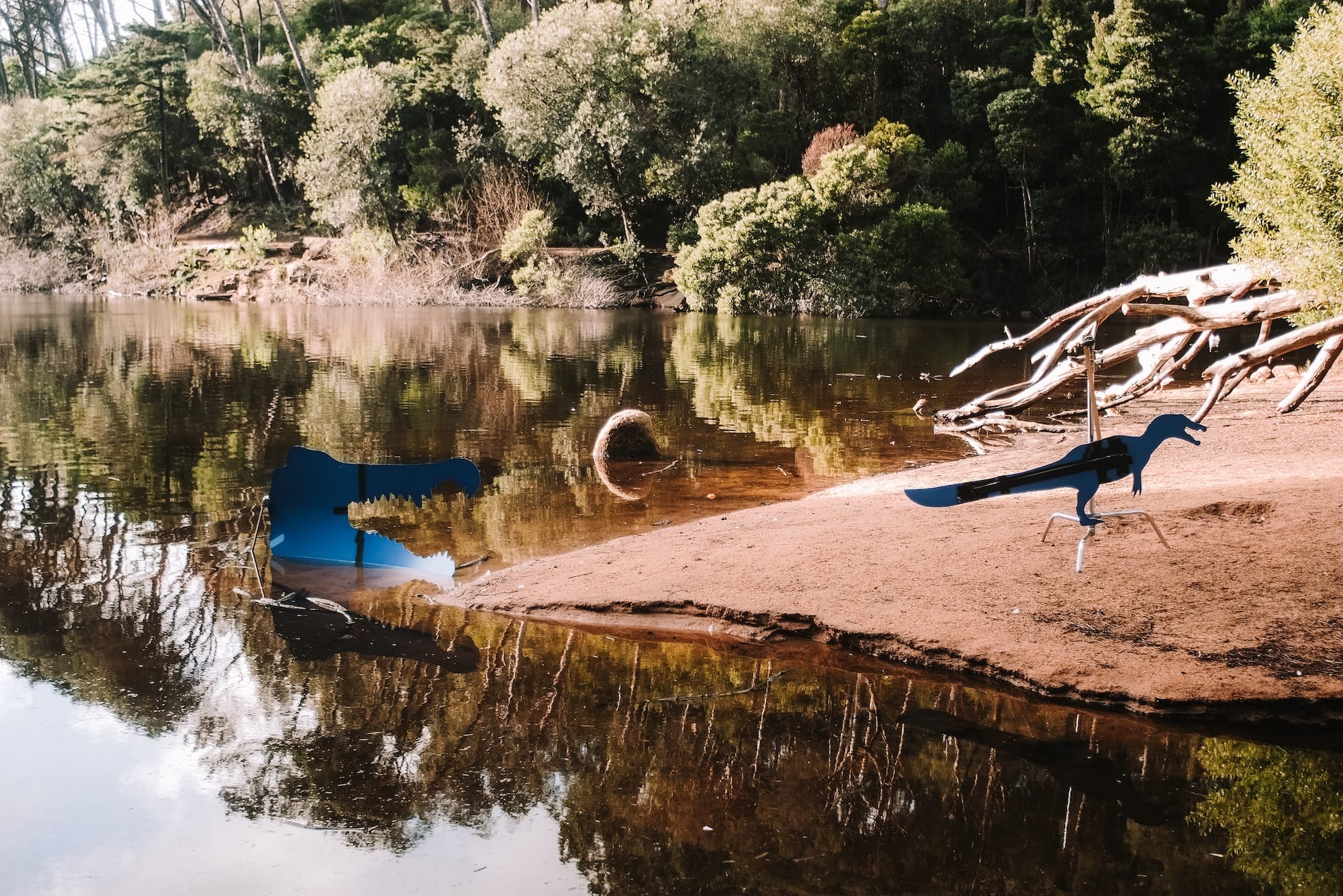 Two blue metal dinosaur cutout sculptures sit on a sandy riverbank surrounded by trees and calm reflective water.