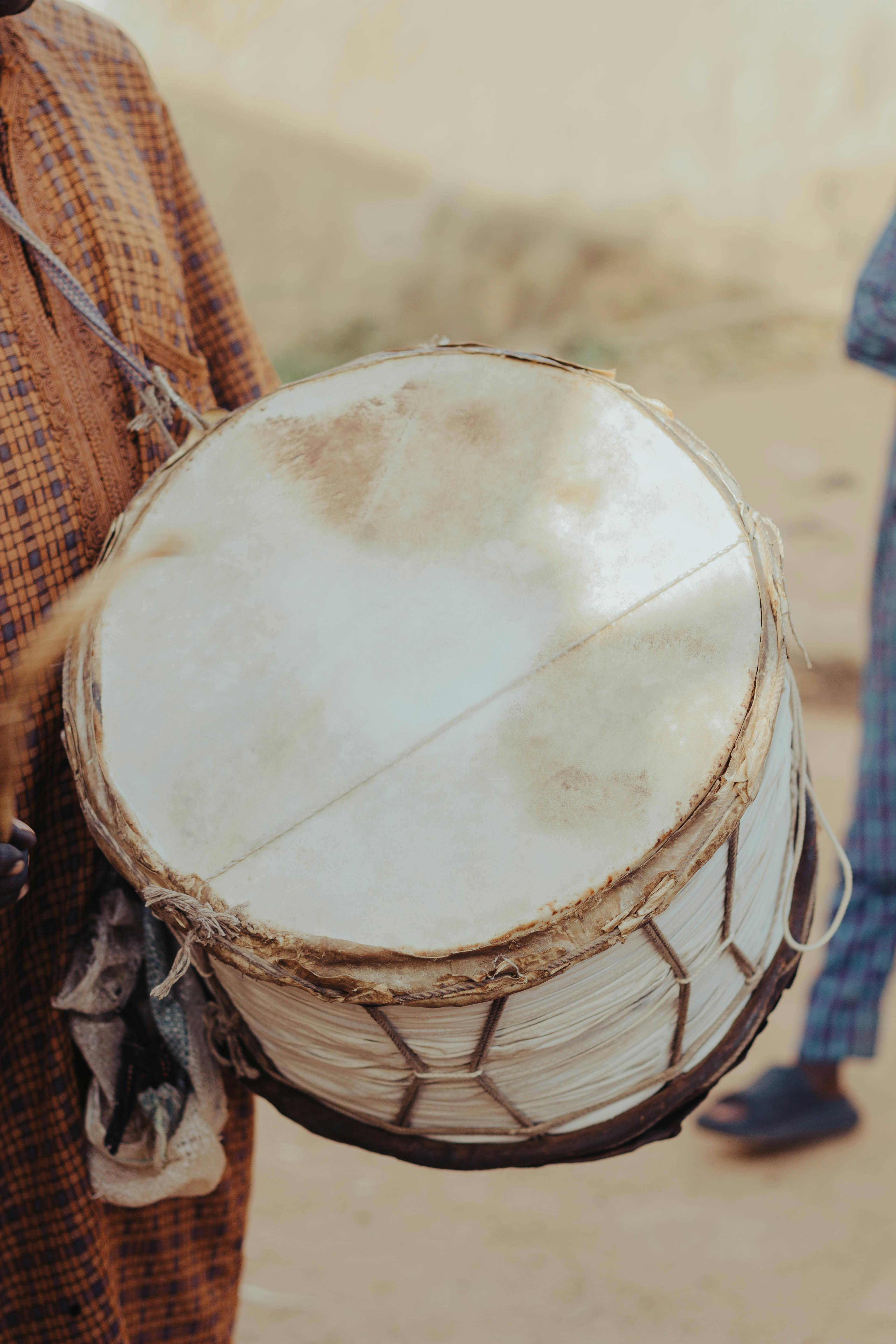 A person in a brown patterned robe holds a large traditional hand drum with a white animal-skin head.