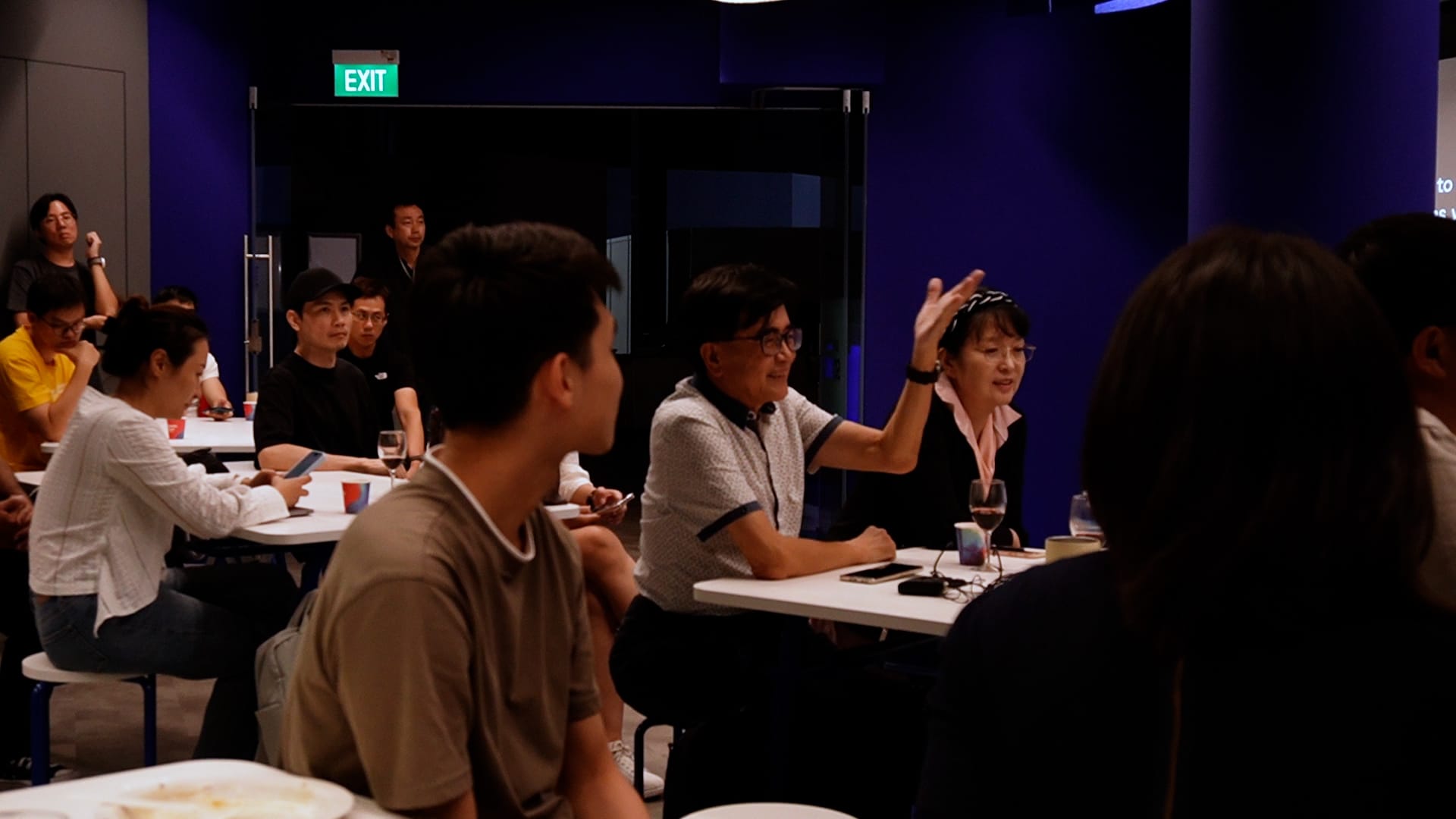A group of people sit at tables in a dark blue room during a meeting, with one woman raising her hand.