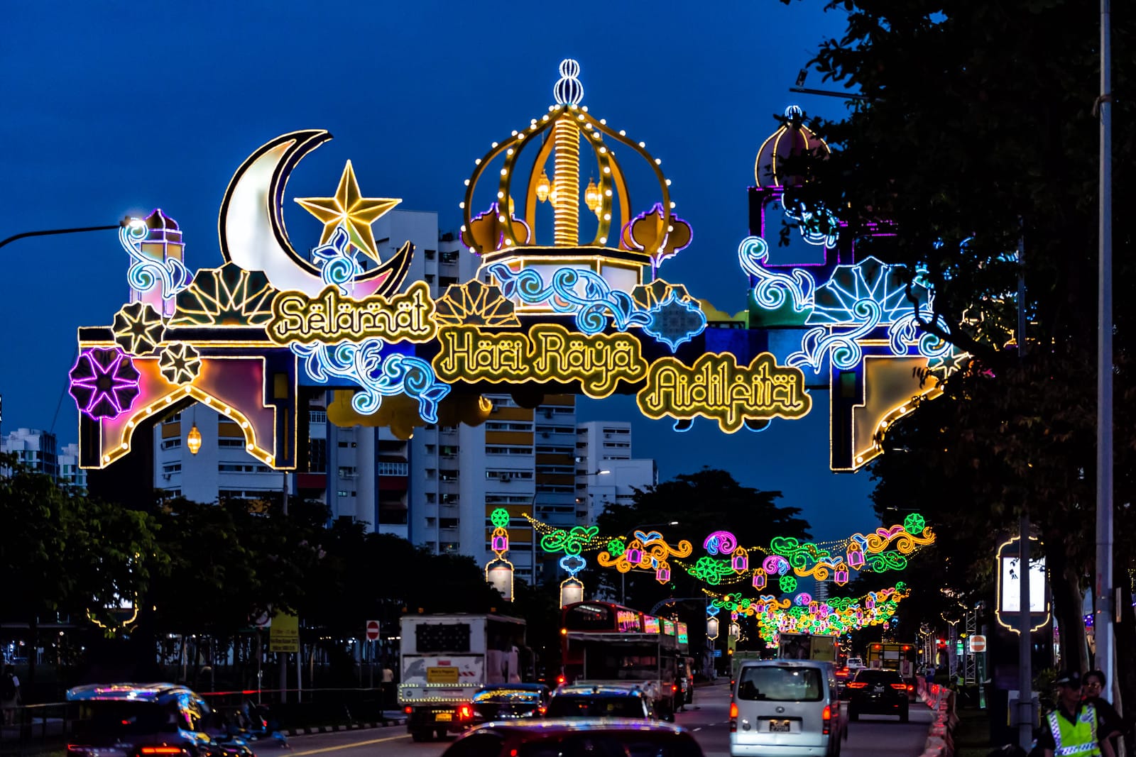 Colorful neon arch reading "Selamat Hari Raya Aidilfitri" spans a busy street at night with festive lights.