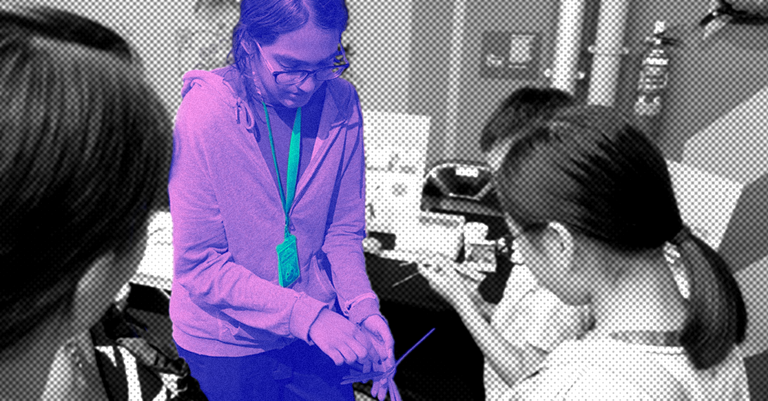 A woman with glasses and a lanyard leans over, demonstrating something to students in a classroom setting.
