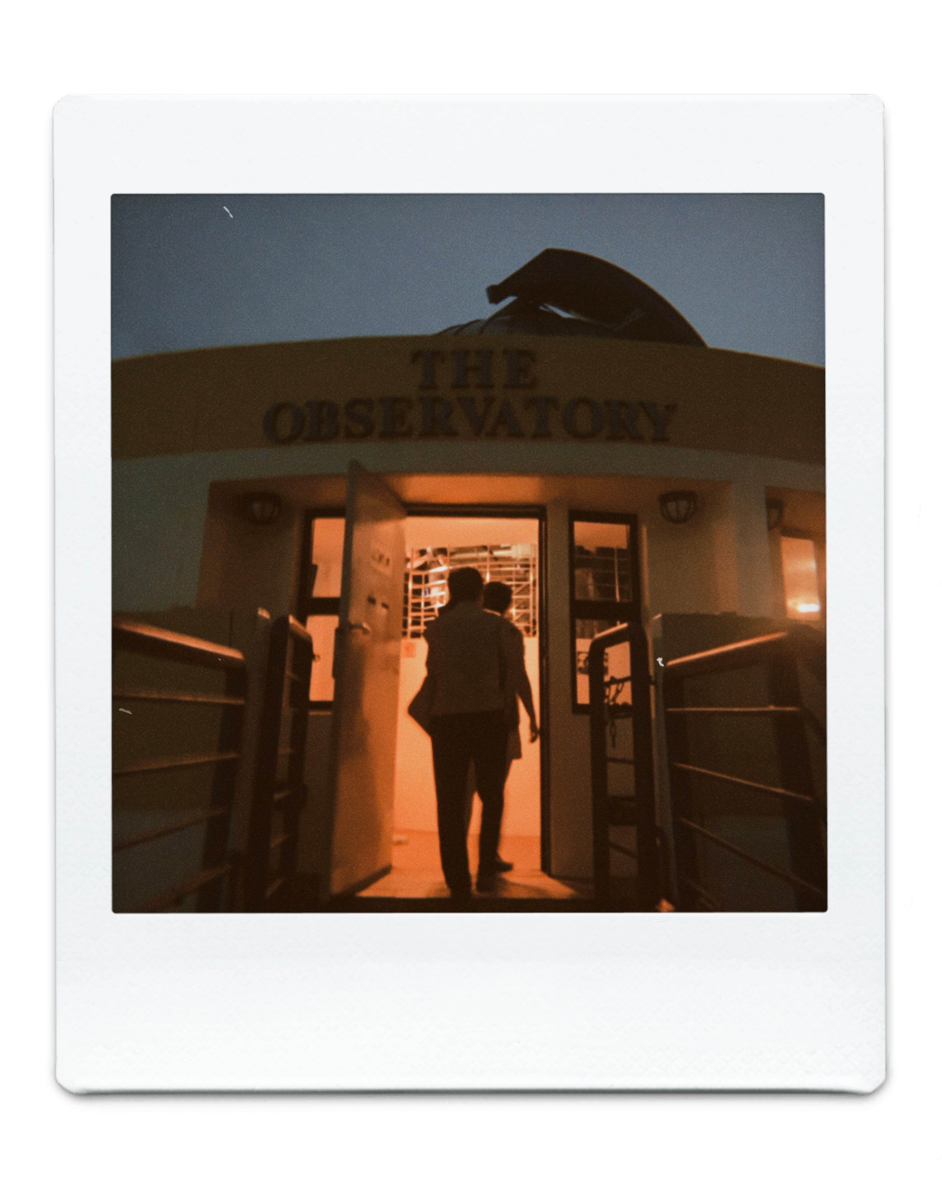 A person's silhouette enters a warmly lit doorway beneath a sign reading "The Observatory" at dusk.